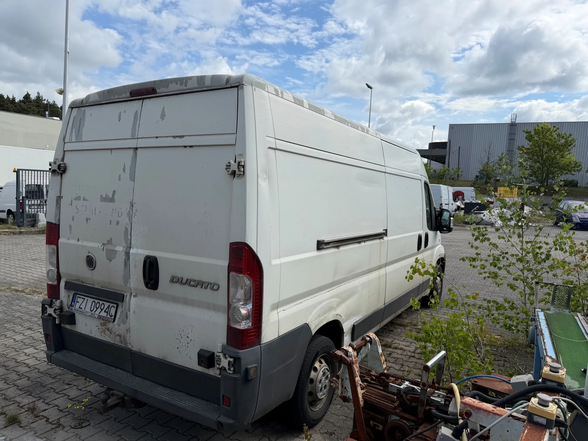 Biały furgon marki Ducato z widocznym numerem rejestracyjnym FZI 0994C, stojący na brukowanym placu. Furgon ma zarysowane ślady zużycia i obłupionej farby na tylnych drzwiach. W tle widoczne są inne pojazdy, przemysłowe budynki, drzewa oraz niebo z chmurami. Na pierwszym planie po prawej stronie znajdują się części maszyn i zielony pas. Tekst na furgonie: DUCATO.