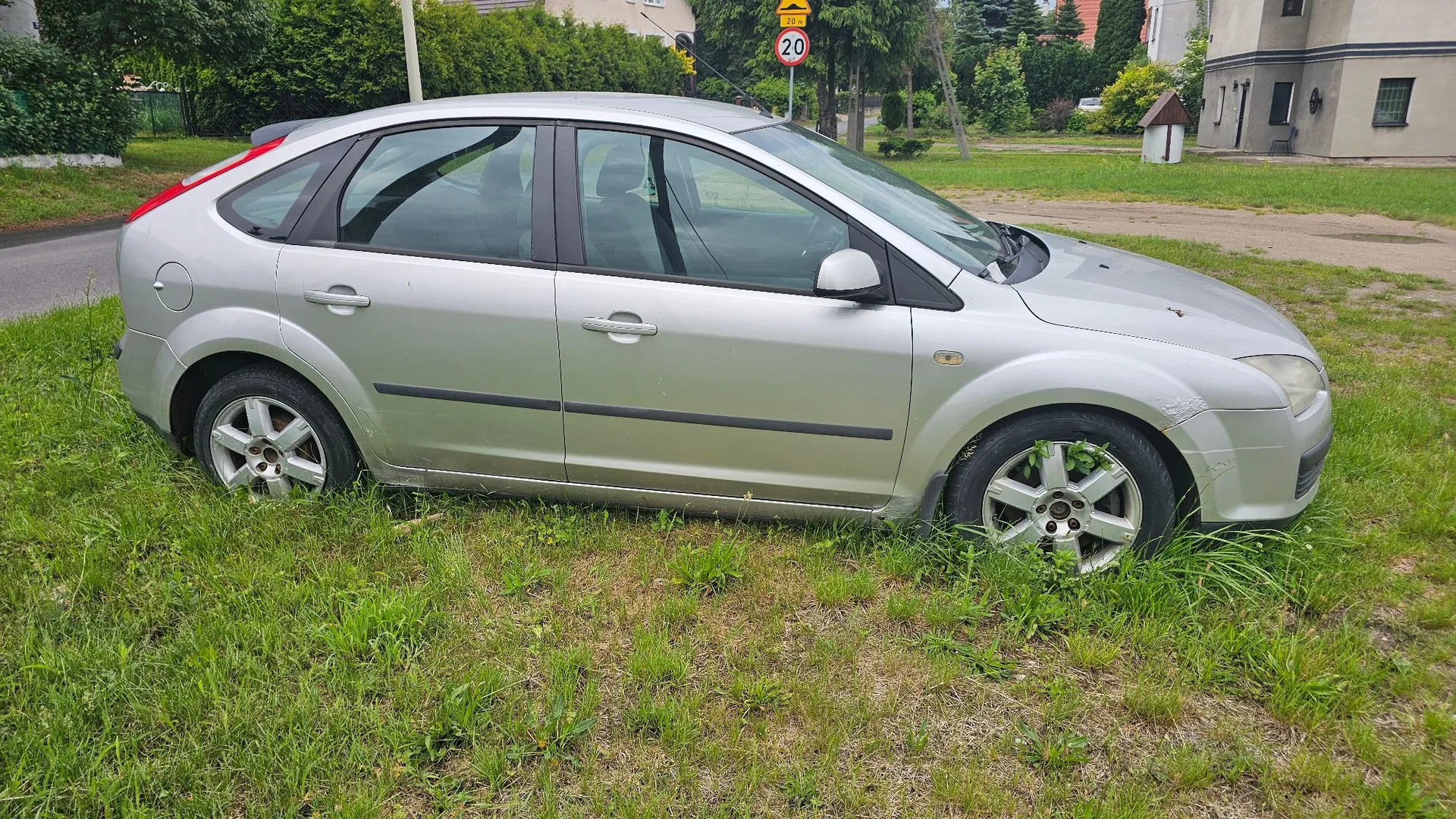 Srebrny samochód osobowy typu hatchback zaparkowany na trawie. Pojazd jest widoczny z profilu od strony pasażera. W tle widać zielone drzewa, krzewy oraz fragment budynku i znak drogowy z ograniczeniem prędkości do 20 km/h.