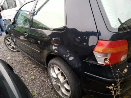 Czarny samochód osobowy typu hatchback widoczny z tyłu i boku, zaparkowany na podłożu z drobnych kamieni. Widoczne są tylne światła, koła z aluminiowymi felgami oraz fragment innej, częściowo widocznej białej pojazdu po lewej stronie. W tle zielone krzewy i fragment budynku z oknami. Na pierwszym planie sucha roślina.