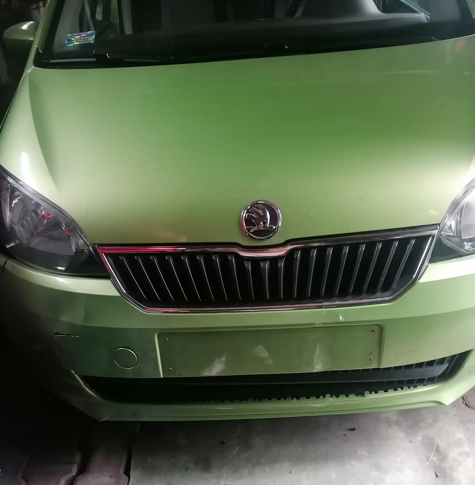 Przednia część zielonego samochodu marki Škoda. Widoczna jest chromowana atrapa chłodnicy z logo marki, reflektory, puste miejsce na tablicę rejestracyjną oraz fragment przedniej szyby z małym naklejką w lewym górnym rogu. Samochód stoi na betonowej powierzchni.
