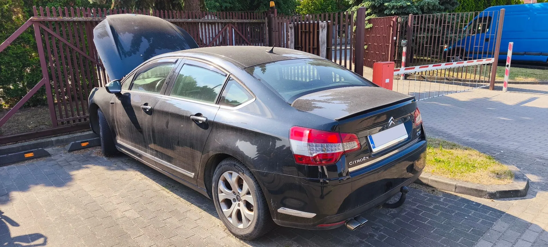 Czarny, zakurzony samochód osobowy marki Citroën zaparkowany tyłem na kostce brukowej. Maski samochodu jest otwarta. Pojazd znajduje się przed brązowym drewnianym ogrodzeniem. W tle widoczny jest niebieski furgon oraz biało-czerwona szlabanowa brama wjazdowa.