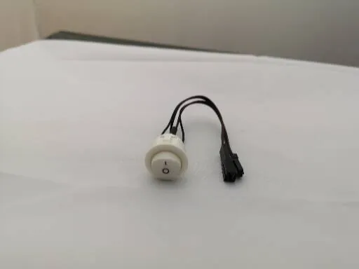 Mały, biały przełącznik okrągły z czarnym symbolem zasilania, połączony z czarnymi przewodami elektrycznymi zakończonymi czarną wtyczką, leżący na białym tle.