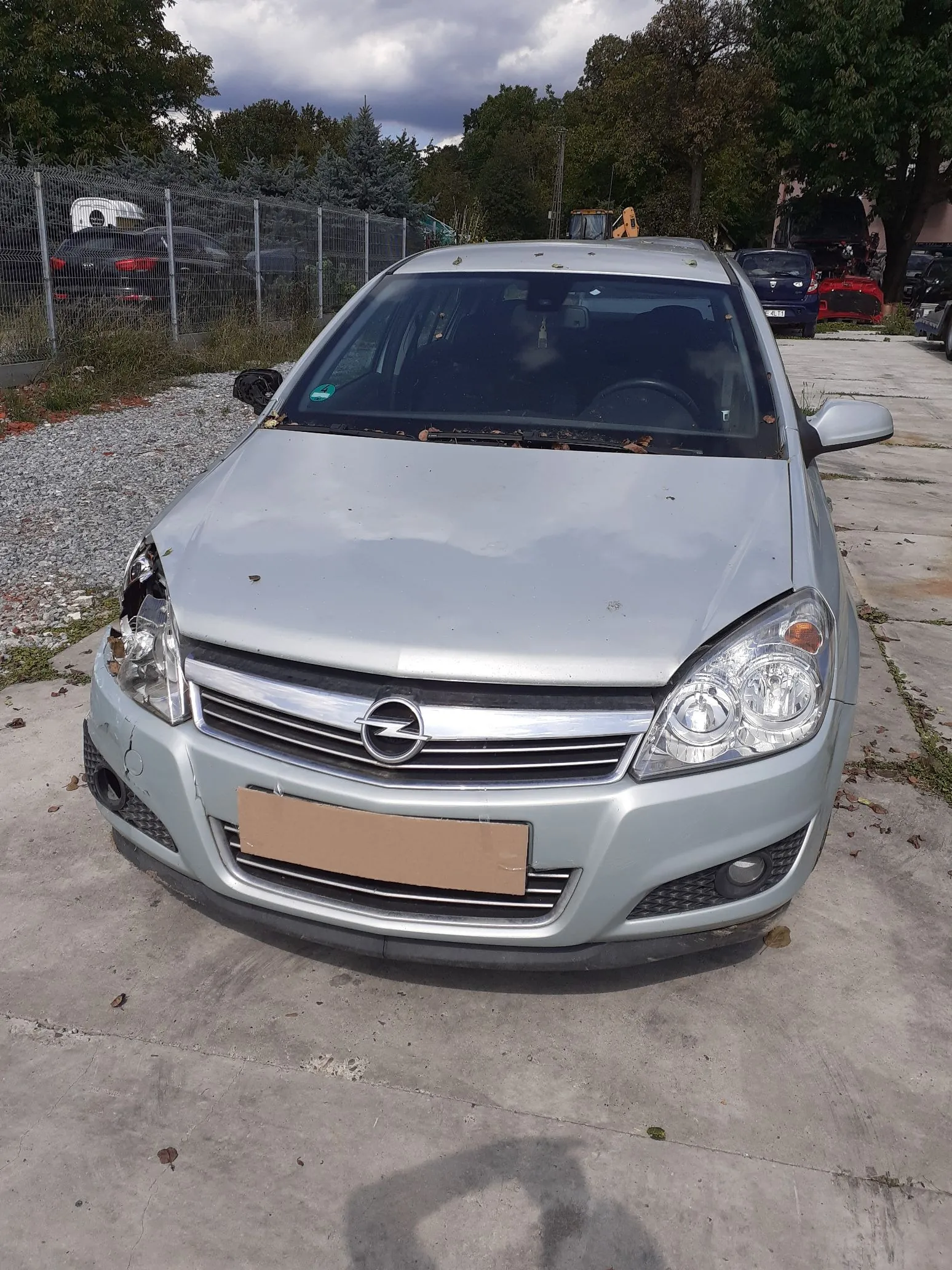 Srebrny samochód marki Opel zaparkowany na betonowym podłożu. Pojazd jest uszkodzony w przedniej części po lewej stronie, gdzie brakuje fragmentu klosza reflektora i widoczne są zniszczenia zderzaka. Na miejscu tablicy rejestracyjnej znajduje się kawałek brązowego kartonu. W tle widoczny jest ogrodzony parking z innymi samochodami oraz drzewa pod zachmurzonym niebem.