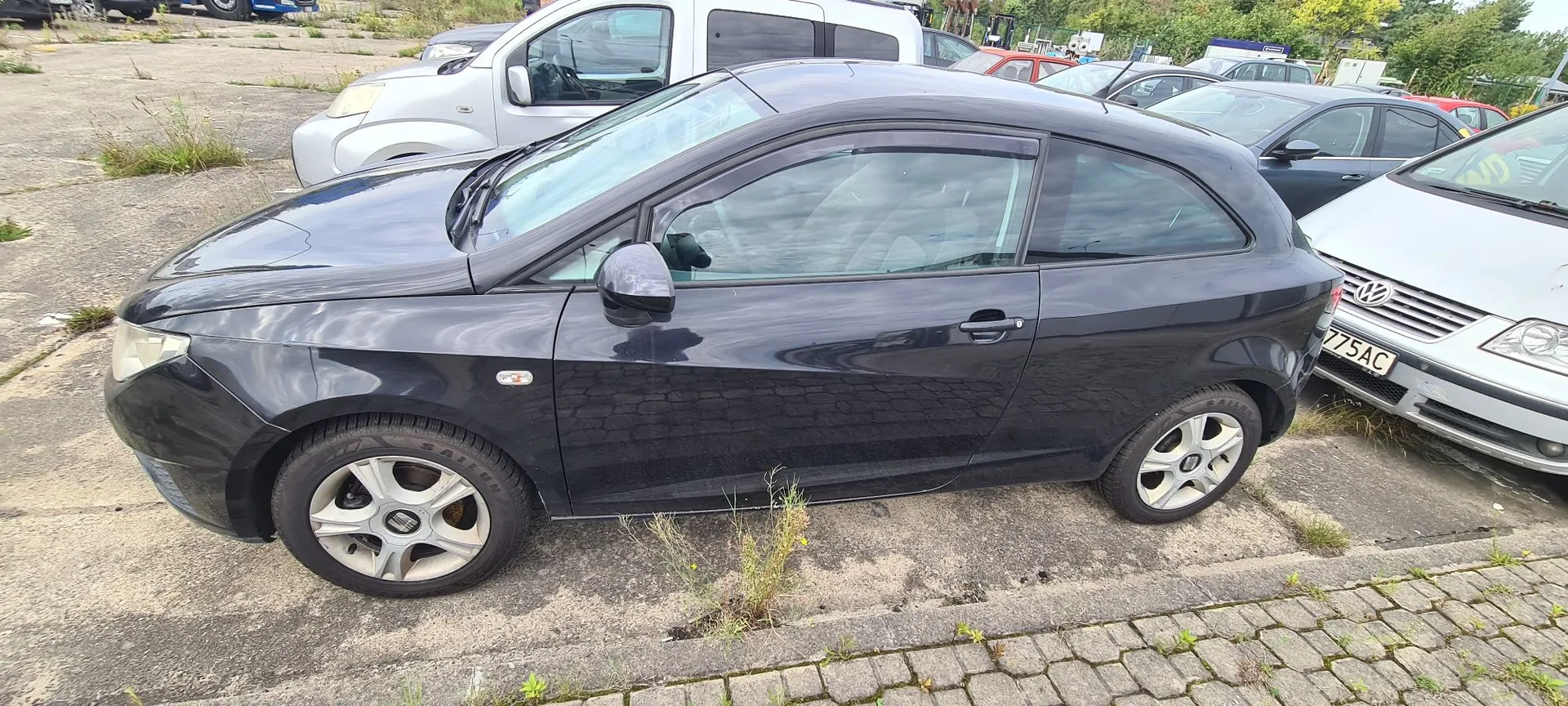 Czarny, trzydrzwiowy samochód osobowy zaparkowany na betonowym placu. Auto jest pokazane z profilu od strony kierowcy. W tle widoczne są inne zaparkowane pojazdy, w tym srebrny van oraz biały samochód po prawej stronie z widoczną częścią tablicy rejestracyjnej o treści '775AC'. Podłoże to szary beton z kępkami trawy i kostką brukarską na pierwszym planie.