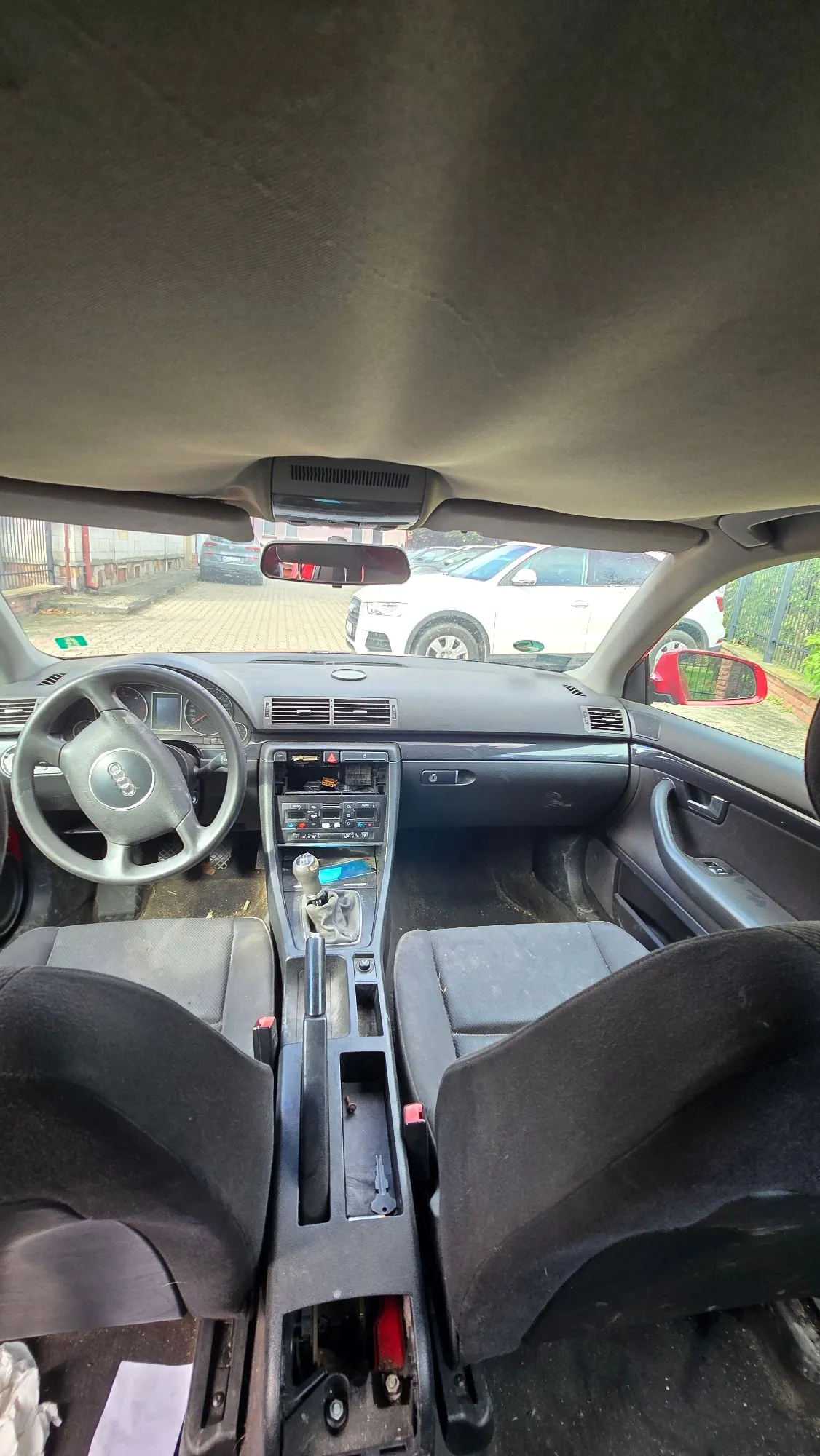 Wnętrze samochodu widziane z perspektywy tylnego siedzenia. Widoczna jest deska rozdzielcza, kierownica z logo Audi, lewy i prawy przedni fotel z czarnej tkaniny oraz konsola środkowa z drążkiem zmiany biegów. Przez przednią szybę widać biały samochód zaparkowany na zewnątrz oraz fragmenty otoczenia.