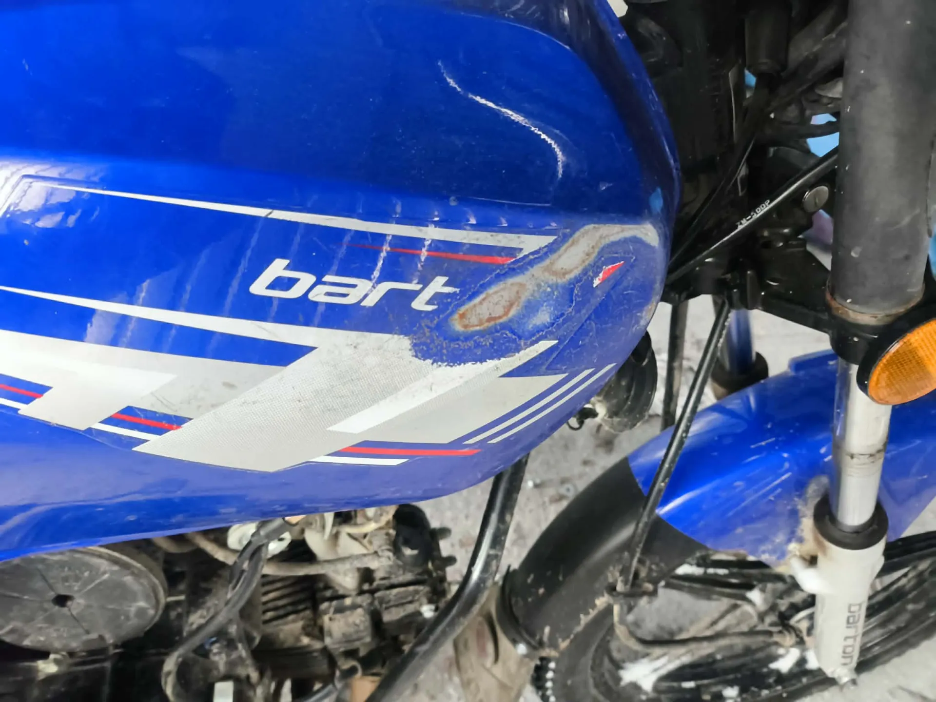 Zbliżenie na niebieski zbiornik paliwa motocykla z białym napisem "bart" oraz białymi i czerwonymi pasami. Na zbiorniku widoczne są zadrapania, uszkodzenia lakieru i plamy rdzy. W tle części mechaniczne motocykla, czarna widelec przedni z napisem "Dafon" oraz pomarańczowy kierunkowskaz. Podłoże betonowe z widocznymi śladami brudu.