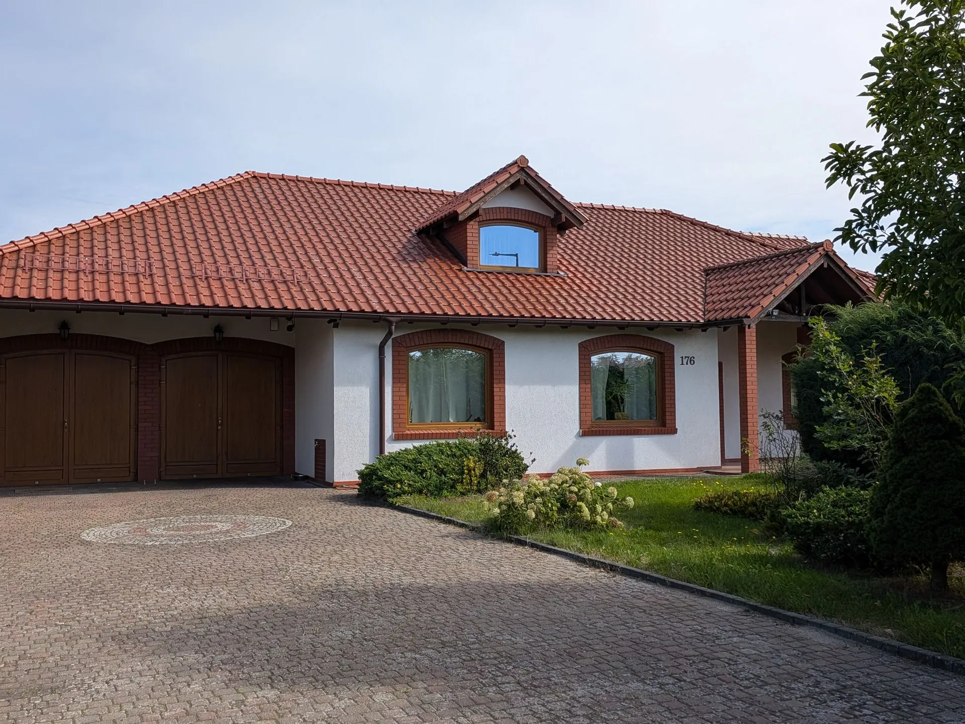 Jednokondygnacyjny dom z poddaszem, kryty czerwonymą dachówką ceramiczną. Budynek ma białe ściany z ceglanymi obramowaniami wokół okien i bram garażowych. Po lewej stronie znajdują się dwie brązowe bramy garażowe. Na fasadzie widoczne są dwa łukowate okna oraz numer domu '176'. Przed domem znajduje się podjazd z szarej kostki brukowej z dekoracyjnym, okrągłym wzorem. Wzdłuż domu rosną niskie krzewy i trawnik, a po prawej stronie widoczne są gęste, zielone drzewa i krzewy.