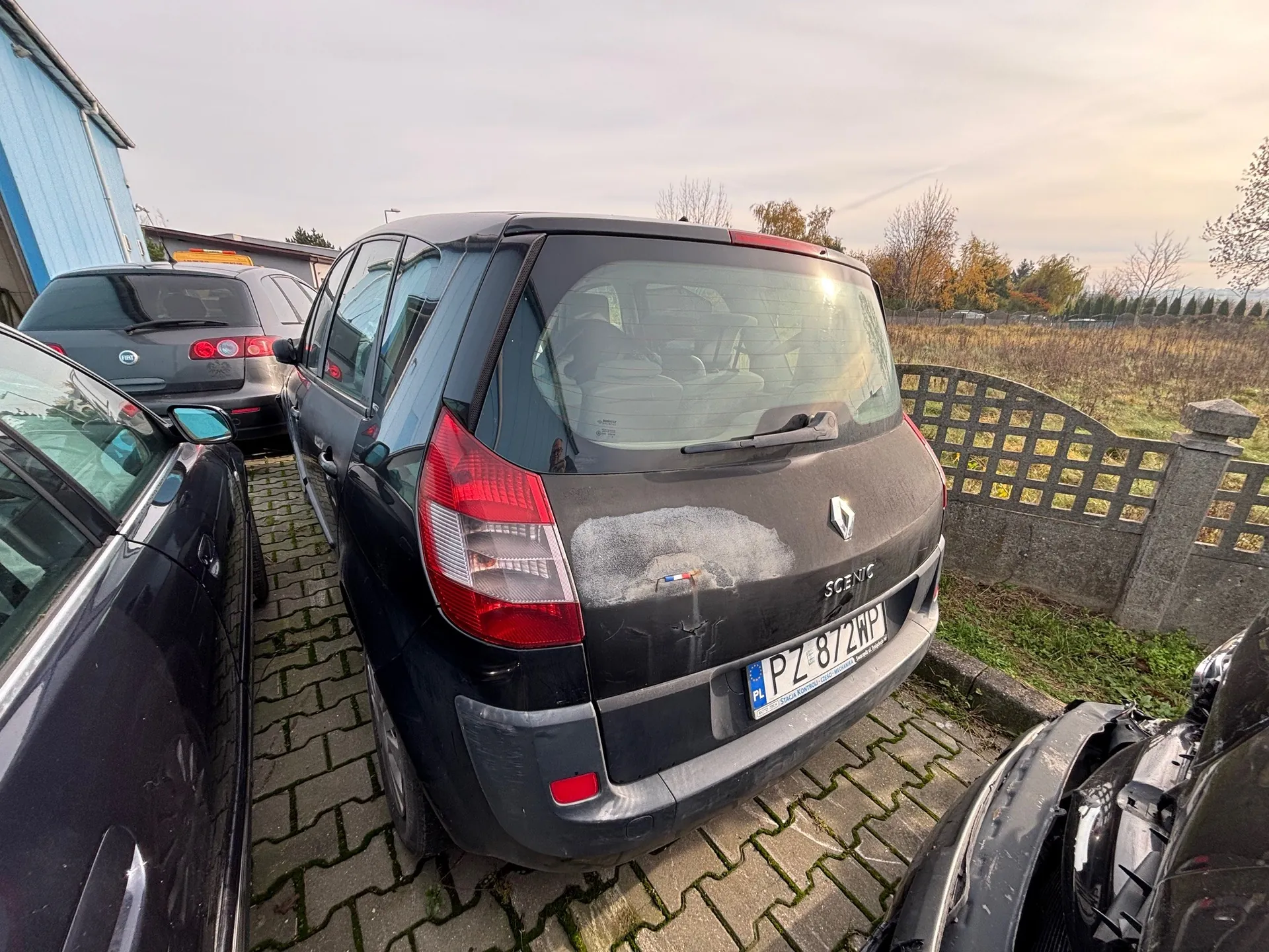 Czarny samochód Renault Scenic zaparkowany tyłem do obserwatora na brukowanej powierzchni. Na tylnej szybie widoczny jest wycieraczka, a na bagażniku napis "SCENIC" oraz tablica rejestracyjna "PZ 872WP" z flagą UE i oznaczeniem "PL". Po lewej stronie częściowo widoczny czarny samochód, za Renault szary Fiat z żółtym znakiem taksówki na dachu. W tle niebieski budynek, kamienny płot z siatką, suche trawy, drzewa z jesiennymi liśćmi i pochmurne niebo. Na bagażniku Renault widoczne są ślady uszkodzenia i biała plama.