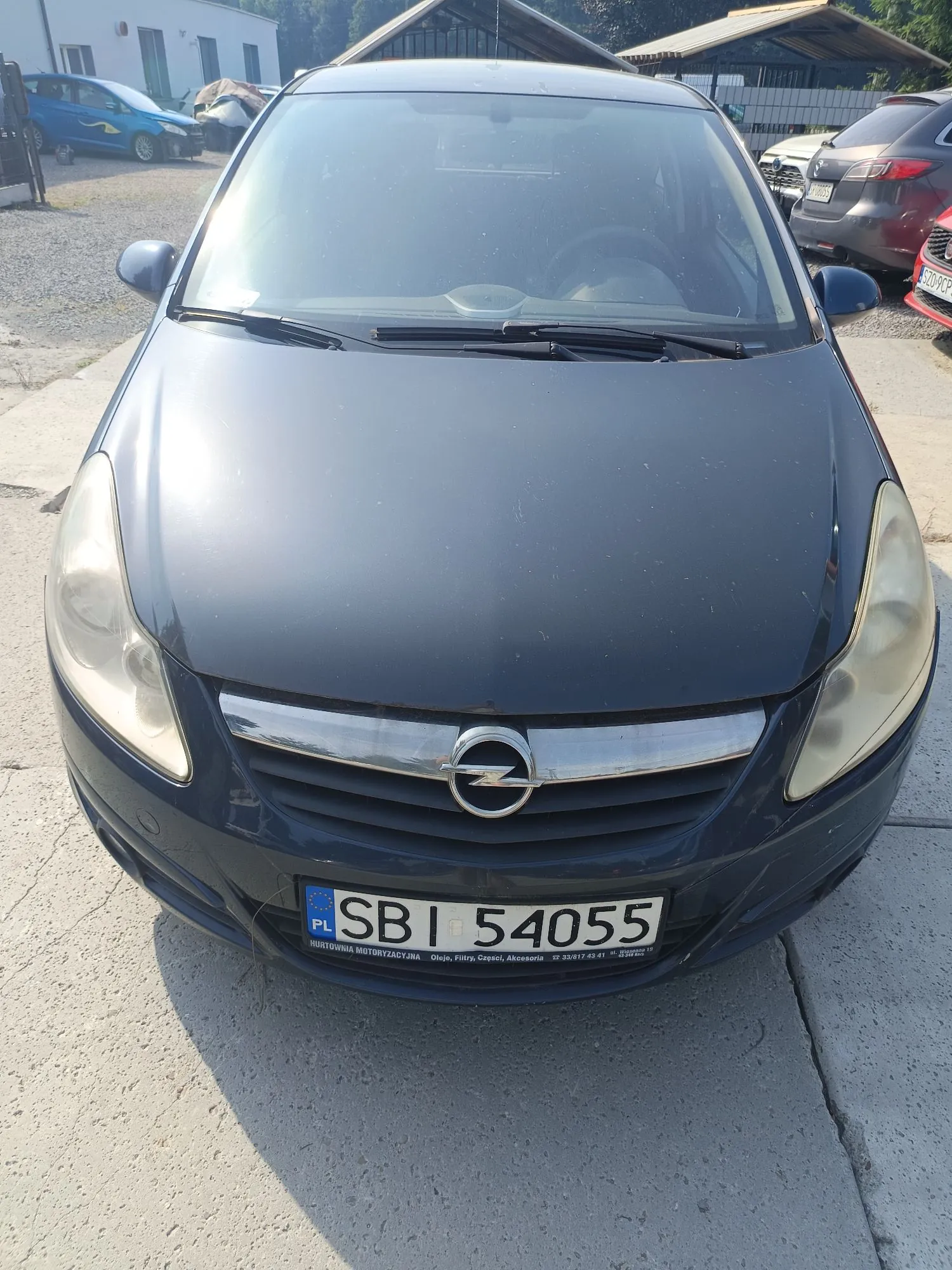 Zdjęcie przodu ciemnoniebieskiego samochodu marki Opel, zaparkowanego na betonowym podłożu. Widoczna jest maska, przednia szyba oraz grill z logo producenta. W tle znajdują się inne samochody oraz budynki.