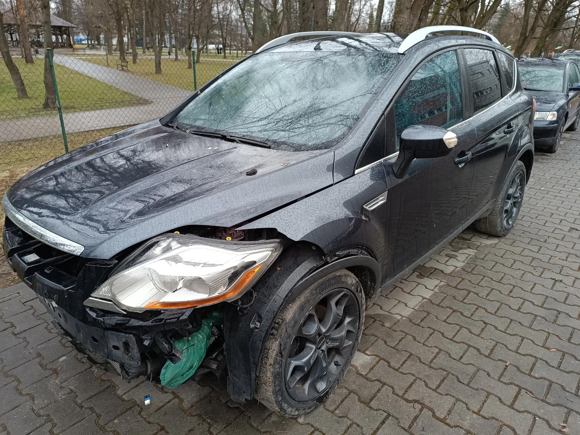 Czarny SUV z uszkodzonym przodem zaparkowany na brukowanej powierzchni. Zdeformowana maska, brak przedniego zderzaka, widoczne uszkodzenia w okolicach reflektora i pasa przedniego. Przy lewym przednim kole znajduje się zielona torba. W tle siatkowe ogrodzenie, trawnik, drzewa bez liści, chodnik oraz drugi czarny samochód. Powierzchnia pojazdu i otoczenia jest wilgotna, co sugeruje niedawny deszcz lub mżawkę. Niebo jest pochmurne.