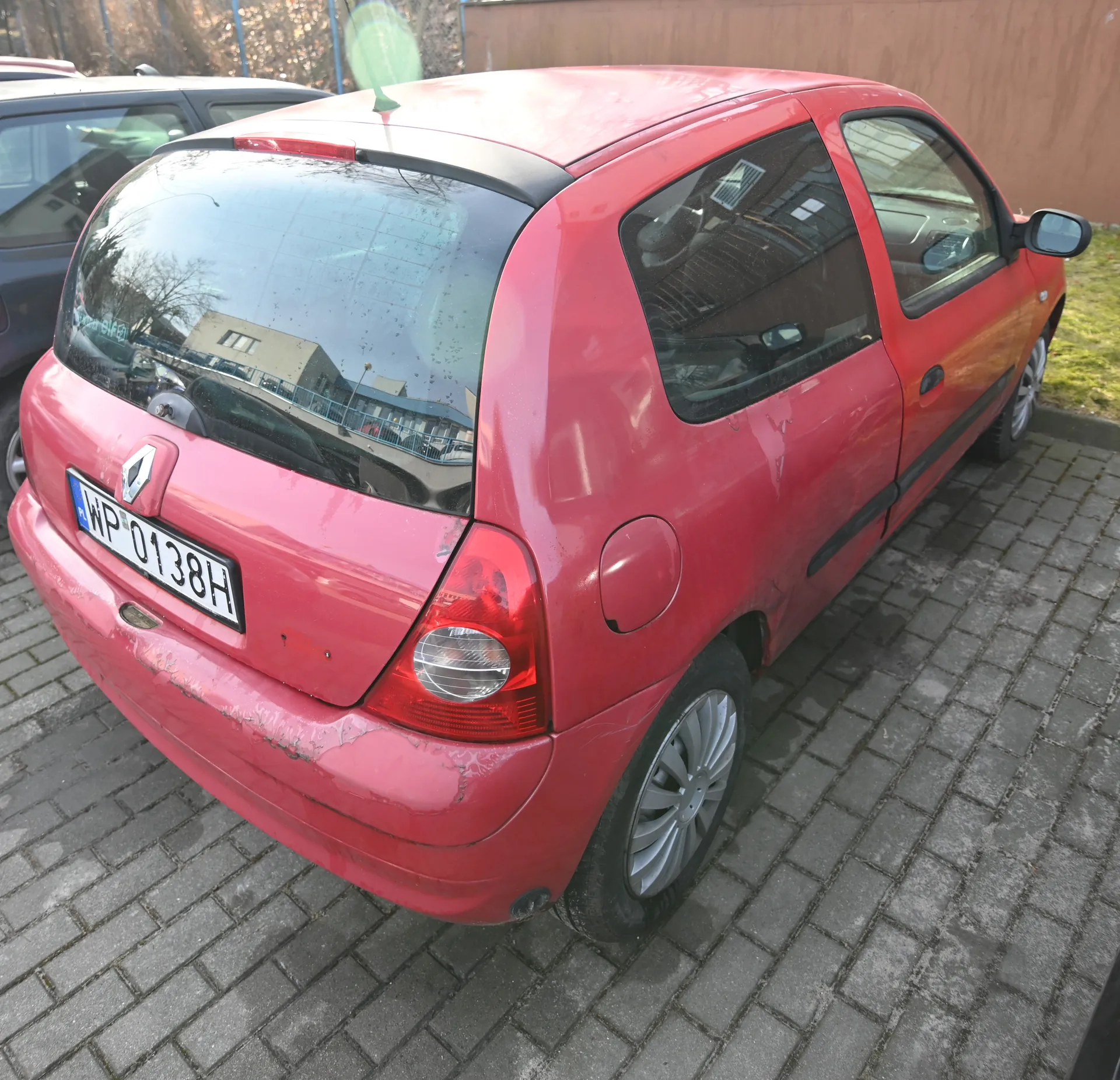 Czerwony samochód osobowy marki Renault zaparkowany na brukowanej powierzchni. Widoczny tył pojazdu z tablicą rejestracyjną WP 0138H. W tle drugi samochód, drewniana płotowa konstrukcja oraz trawa. Na szybie tylnej odbicie budynku i drzew.