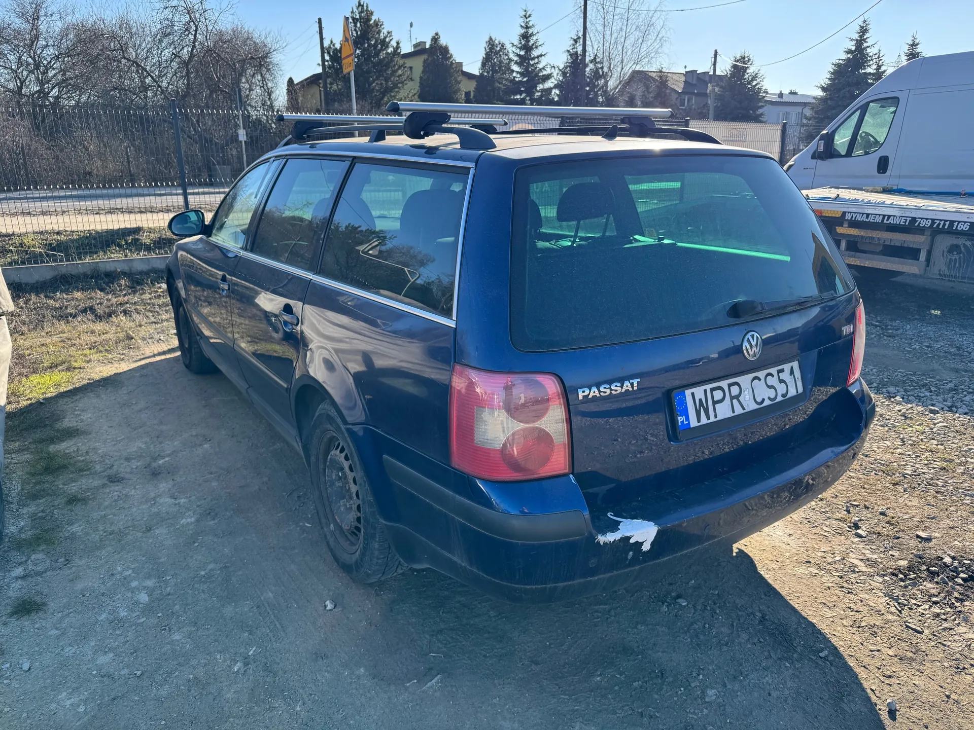 Ciemnoniebieski samochód osobowy typu kombi marki Volkswagen Passat z tablicą rejestracyjną WPR CS51 (PL) zaparkowany na żwirowym podjeździe. Na tylnym zderzaku widoczne uszkodzenie z białą farbą. Samochód wyposażony w dachowy bagażnik. W tle biały furgon z napisem WYNAJEM LAWET 799 711 166, metalowe ogrodzenie, suche drzewa, domy i drogowskaz. Niebo jest bezchmurne.