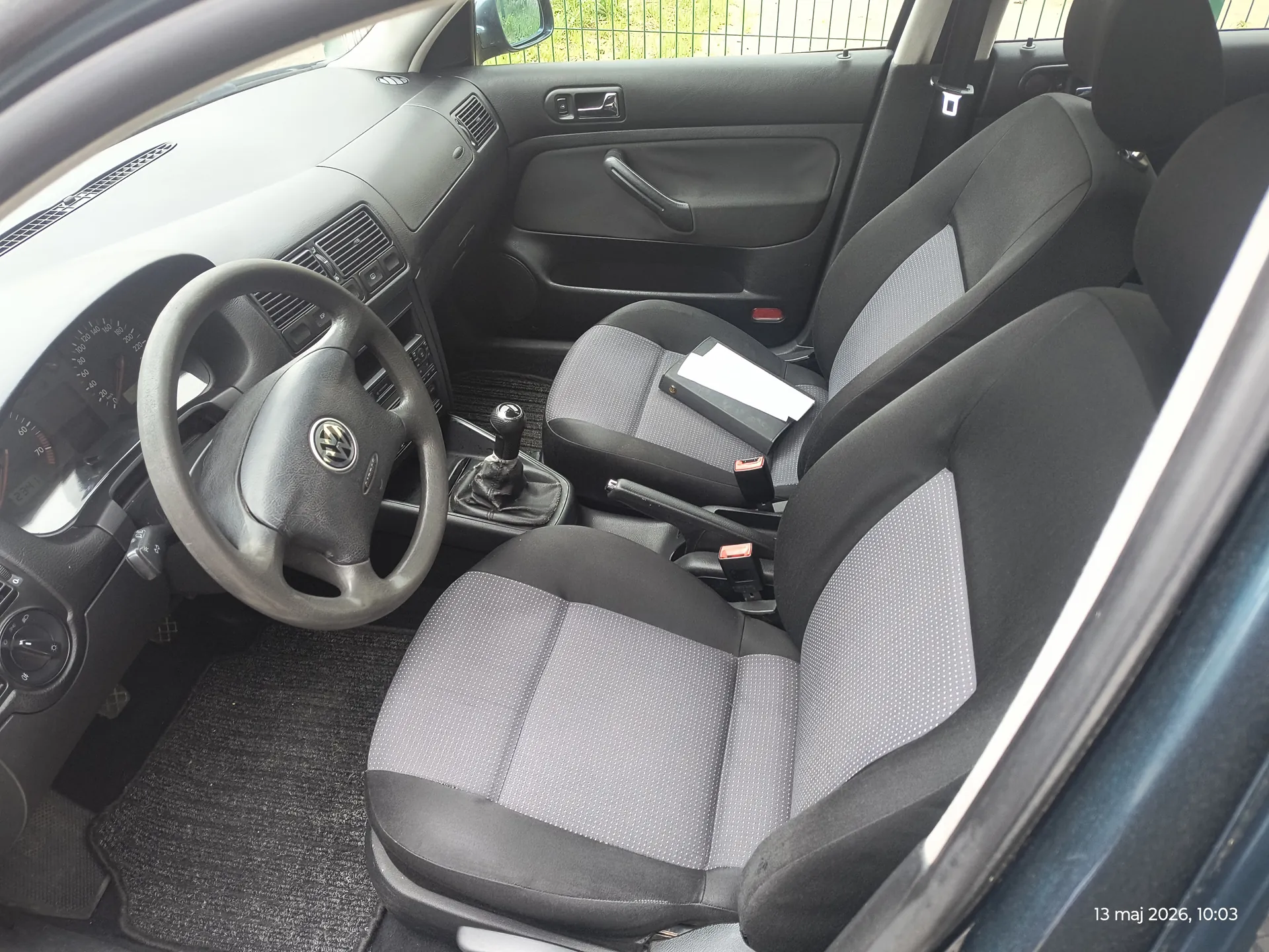 Wnętrze samochodu marki Volkswagen z czarną tapicerką i siedzeniami w szaro-czarną kropkę. Widoczna kierownica z logo VW, deska rozdzielcza z licznikami, dźwignia zmiany biegów, drzwi z uchwytem i sterowaniem okien. Na siedzeniu pasażera leży czarna książka lub dokument. W prawym dolnym rogu obrazu widoczny tekst: 13 maj 2026, 10:03. W tle przez okno widać siatkę ogrodzeniową i zieleń.