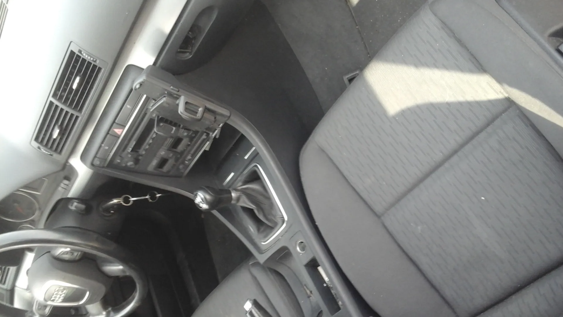 Wnętrze samochodu z widoczną kierownicą z logo Audi, deską rozdzielczą z wentylacją, centralną konsołą z odtwarzaczem i przyciskami, dźwignią zmiany biegów oraz czarnymi siedzeniami tapicerowanymi tkaniną. Kluczyki w stacyjce.