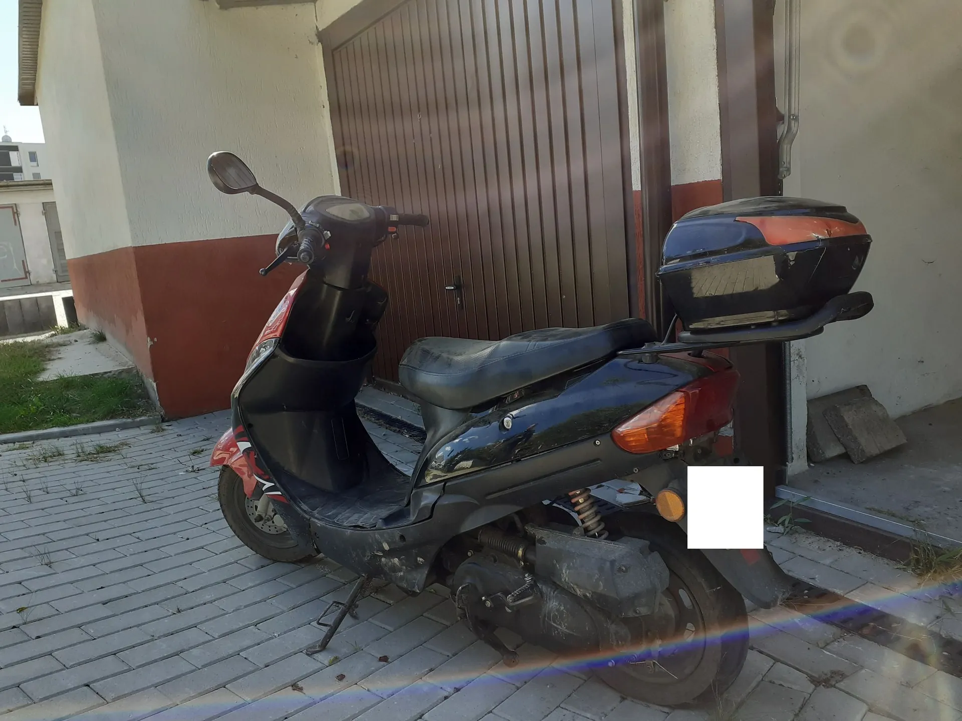Czarny skuter zaparkowany na kostce brukowej przed brązową bramą garażową. Pojazd posiada zamontowany czarny kufer z pomarańczowym elementem na górze. Skuter stoi na bocznej stopce, a jego tył jest skierowany w prawą stronę obrazu. Tablica rejestracyjna jest zakryta białym prostokątem.