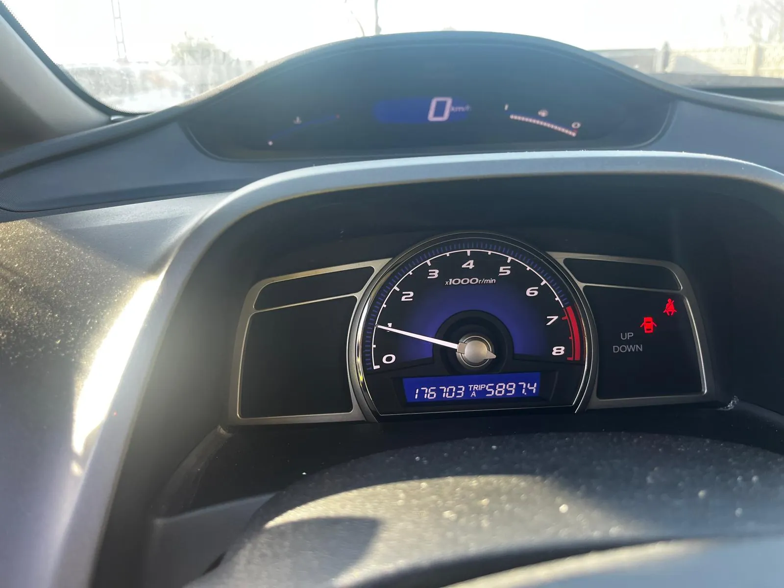 Wnętrze samochodu z widocznym deskiem rozdzielczą. W centrum znajduje się tachometr z niebieskim tłem, zakresem od 0 do 8 (oznaczonym jako x1000 r/min), wskazówką na 0. Poniżej cyfrowy wyświetlacz z napisem "176703 TRIP A 5897.4". Po prawej stronie dwa czerwone symbole ostrzegawcze (pasa bezpieczeństwa i drzwi) oraz napisy "UP" i "DOWN". W tle widoczny prędkościomierz z wartością 0 km/h oraz fragment przedniej szyby z zewnątrz widocznymi budynkami i słonecznym światłem.
