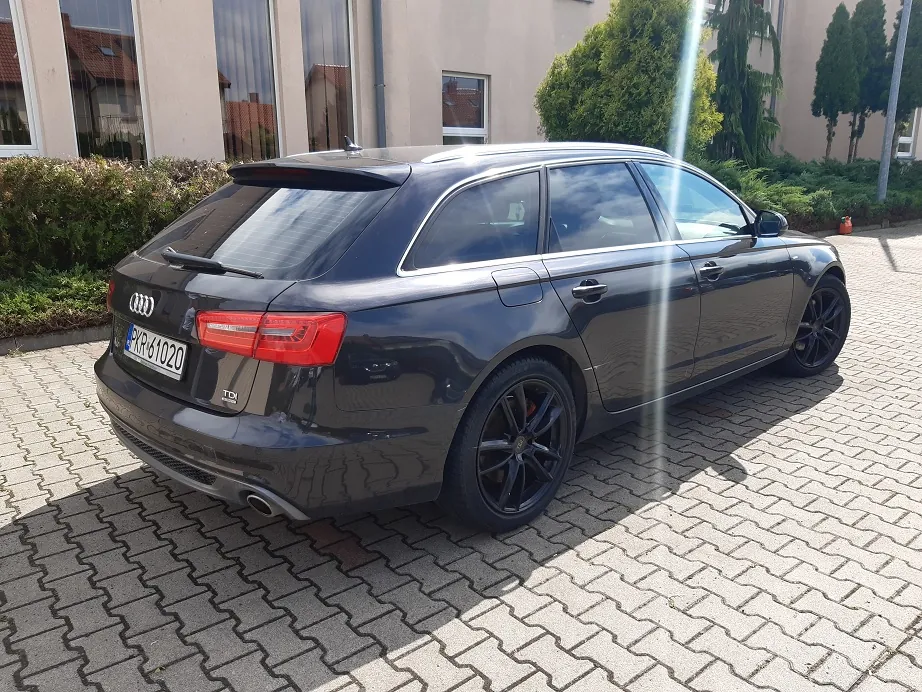 Ciemnoszary samochód marki Audi w nadwoziu kombi, zaparkowany na kostce brukowej przed budynkiem. Widoczny jest tył i prawy bok auta. Samochód ma czarne felgi i polską tablicę rejestracyjną o numerze PKR 61020. W tle widać jasną ścianę budynku z oknami oraz zielone krzewy i drzewa.