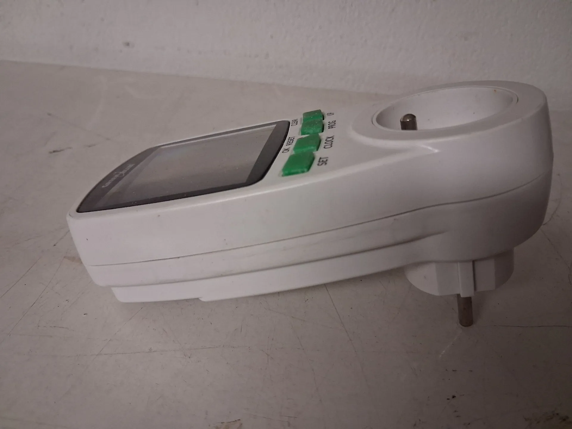 Biały programator czasowy do gniazdka elektrycznego leżący na jasnej powierzchni. Urządzenie posiada ekran LCD, cztery zielone przyciski sterujące z napisami: 'SET', 'CLOCK', 'PROG' oraz 'RESET' oraz gniazdo elektryczne z przodu i wtyczkę z tyłu.