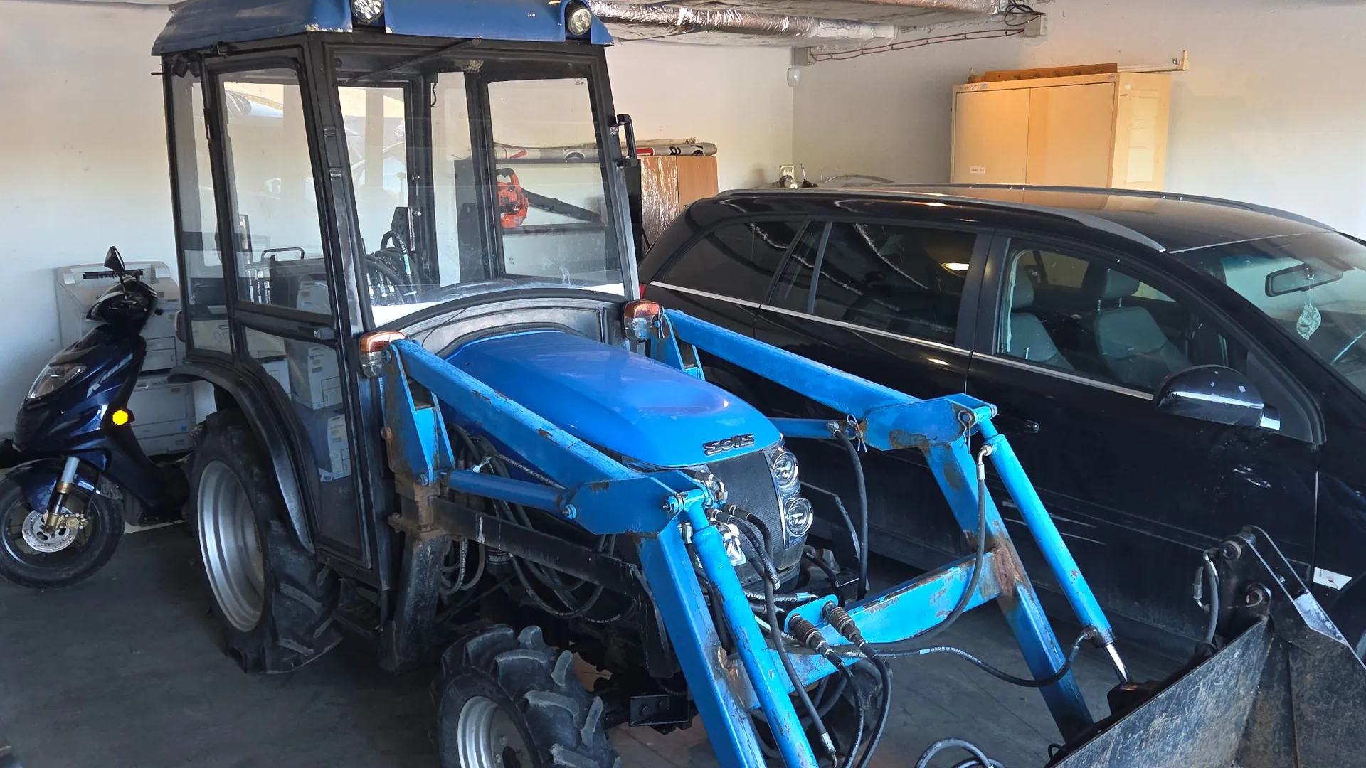 Niebieski traktor z przednim ładowaczem zaparkowany w garażu obok czarnego samochodu osobowego i niebieskiego skutera. Traktor posiada przeszkloną kabinę, a jego ładowacz jest wyposażony w hydrauliczne ramiona. W tle widoczna jest biała ściana oraz drewniana szafka.