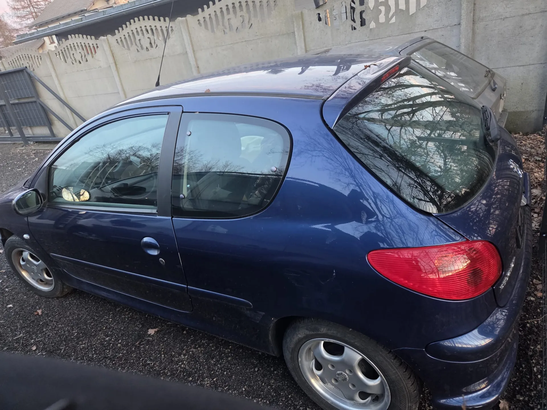 Ciemnoniebieski samochód osobowy typu hatchback zaparkowany na podwórku z żwiru. Widok z boku i tyłu, z widocznymi czerwonymi światłami tylnymi, srebrnymi felgami i czarnymi oknami. W tle betonowy mur z ozdobnymi otworami oraz fragment metalowej bramy. Na ziemi leżą suche liście.