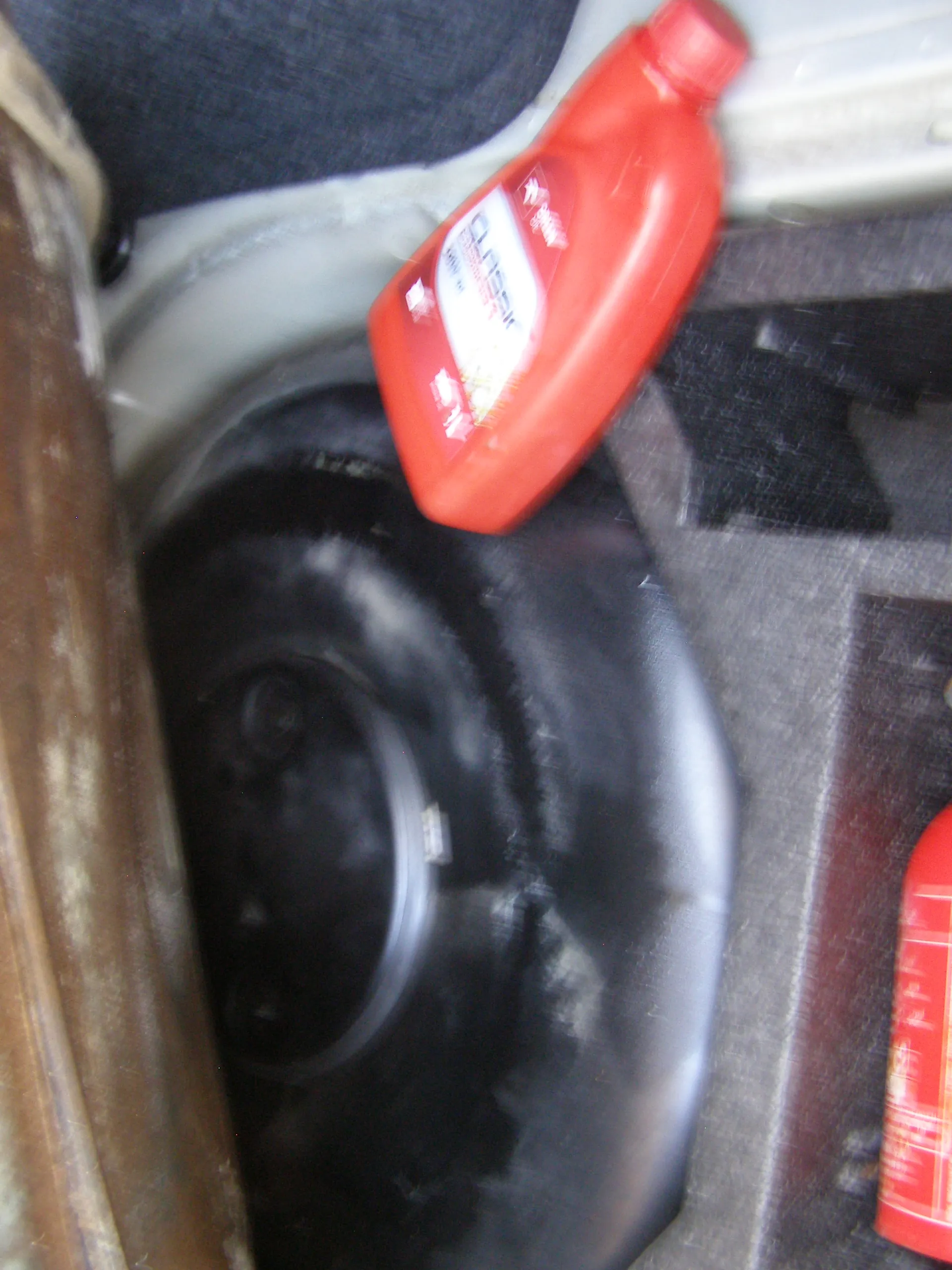 Wnętrze bagażnika samochodu z czarnym kołem zapasowym o metalowym pierścieniu w centrum. Obok znajduje się czerwona butelka z częściowo widocznym napisem 'LIQUID' oraz innym tekstem. W dolnej części obrazu widoczna jest część drugiej czerwonej pojemności. Tło stanowią materiały i elementy metalowe bagażnika.