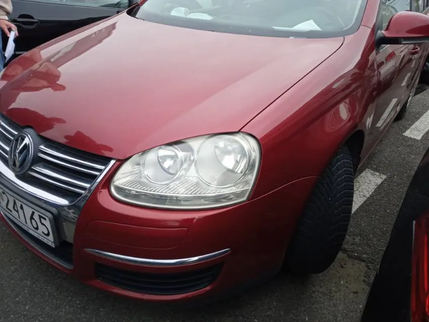 Czerwony samochód marki Volkswagen z przodu, widoczna maska, reflektory, chromowana kratka z logo VW oraz tablica rejestracyjna z częściowo widocznym numerem 24165. Samochód zaparkowany na asfaltowej nawierzchni, w tle fragmenty innych pojazdów.
