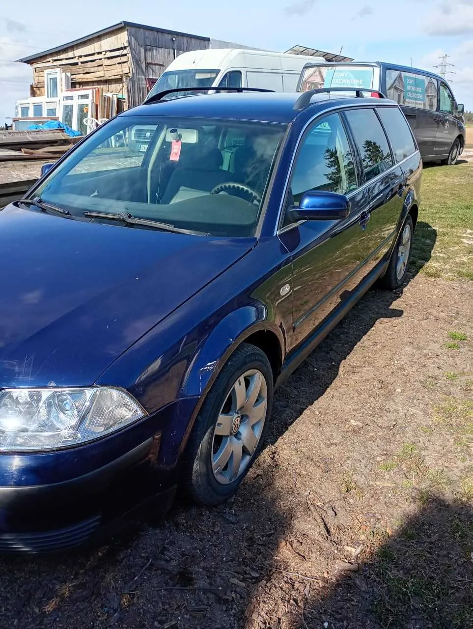 Ciemnoniebieski samochód kombi marki Volkswagen zaparkowany na podwórku z ziemią i trawą. Na przedniej szybie widoczny czerwony znaczek. W tle biały furgon, ciemny van z napisem "Domy Drewniane" oraz drewniana budowla z oknami i materiałami budowlanymi. Jasne niebo z kilkoma chmurami.