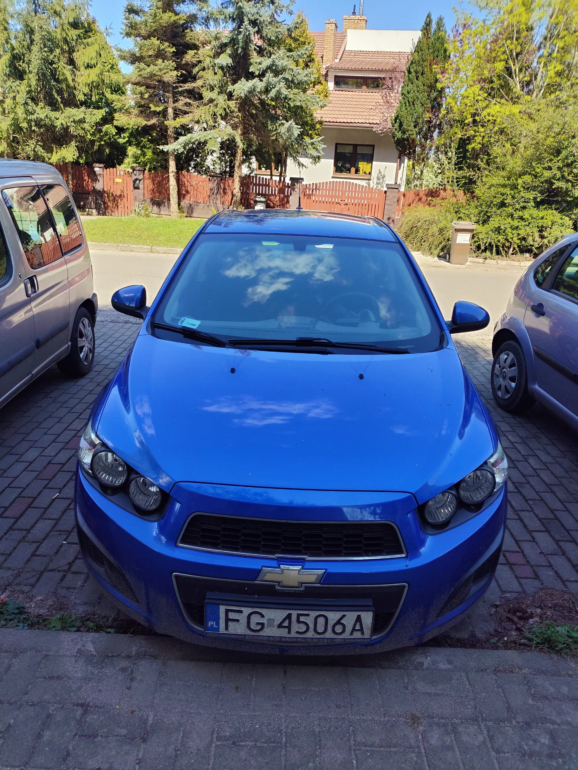 Niebieski samochód marki Chevrolet z tablicą rejestracyjną PL FG 4506A zaparkowany na brukowanym podjeździe. Po obu stronach widoczne są inne pojazdy: srebrny furgon po lewej i szary samochód po prawej. W tle dom z czerwoną dachówką, ogrodzenie z czerwonego drewna, drzewa oraz pojemnik na śmieci. Niebo jest błękitne z kilkoma chmurami.