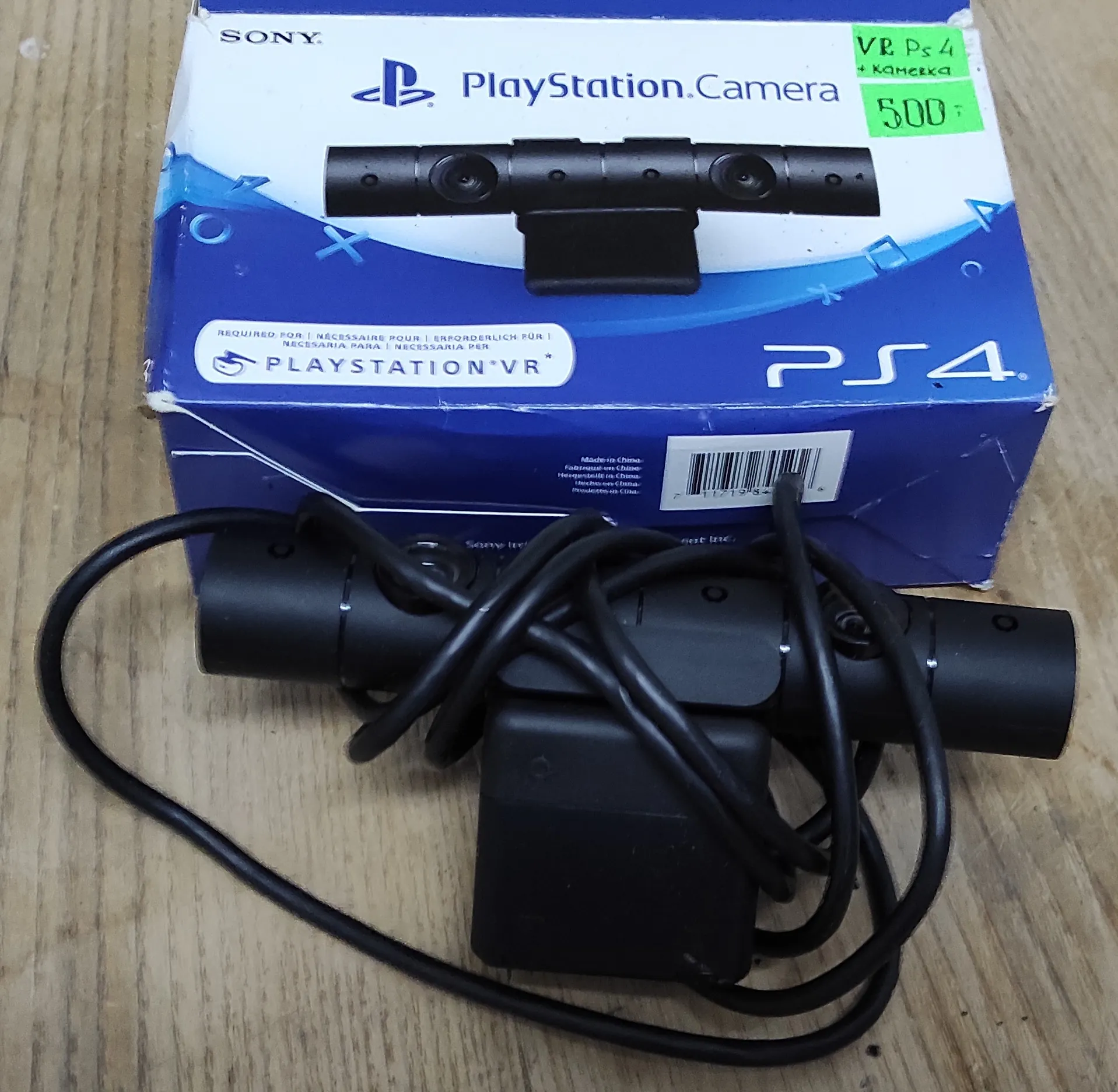 Zdjęcie przedstawiające kamerę PlayStation Camera oraz jej oryginalne opakowanie leżące na drewnianej powierzchni. Pudełko jest niebiesko-białe z logo PlayStation, napisem 'PlayStation Camera' oraz 'PS4'. W prawym górnym rogu pudełka znajduje się zielona naklejka z odręcznym napisem 'VR Ps 4 + камера 500'. Przed pudełkiem leży czarna kamera z podłączonym kablem, który jest częściowo zaplątany wokół urządzenia.
