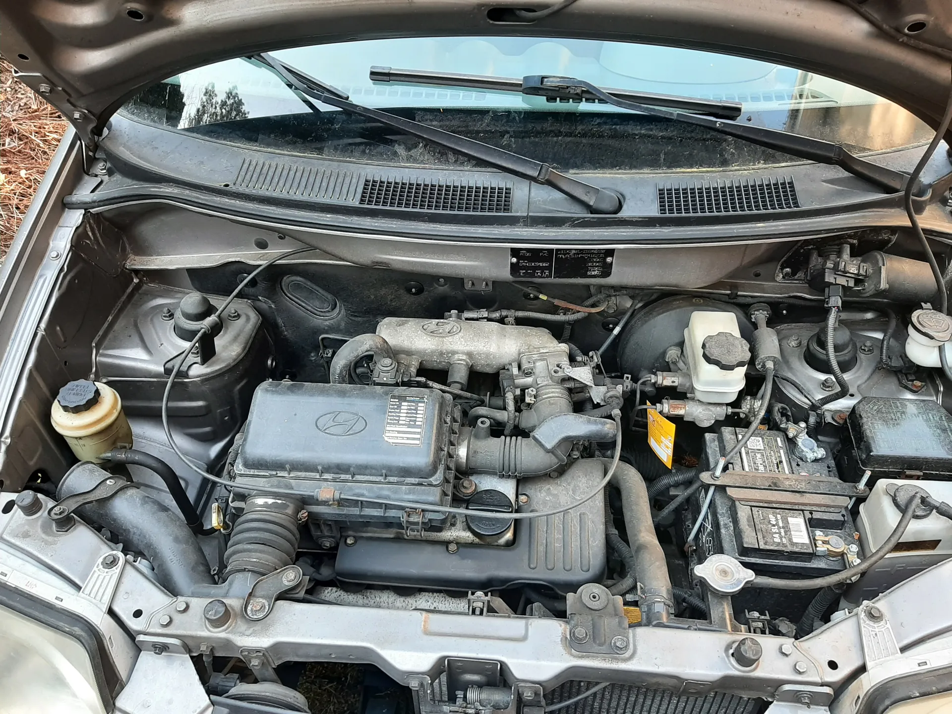 Widok na otwarty maskę samochodu marki Hyundai z widocznym silnikiem. W komorze silnika znajdują się elementy takie jak akumulator po prawej stronie, zbiornik płynu chłodniczego, przewody, rury oraz czarna pokrywa silnika z logo Hyundai. Na pokrywie silnika umieszczona jest tabliczka z nieczytelnym tekstem. W tle widoczne są wycieraczki przednie i przednia szyba. Po lewej stronie obrazu znajduje się żółty zbiornik z czarną nakrętką, a na akumulatorze widoczne są etykiety z kodami. Całość ukazana jest z góry, z otwartym kapotem.