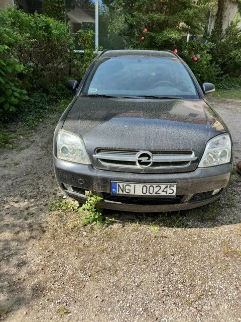 Zdjęcie przodu czarnego samochodu marki Opel, który jest pokryty grubą warstwą kurzu i brudu. Pojazd posiada polską tablicę rejestracyjną o numerze NG1 00245. Samochód stoi na piaszczystym podłożu, w tle widoczna jest gęsta zielona roślinność i fragment budynku.