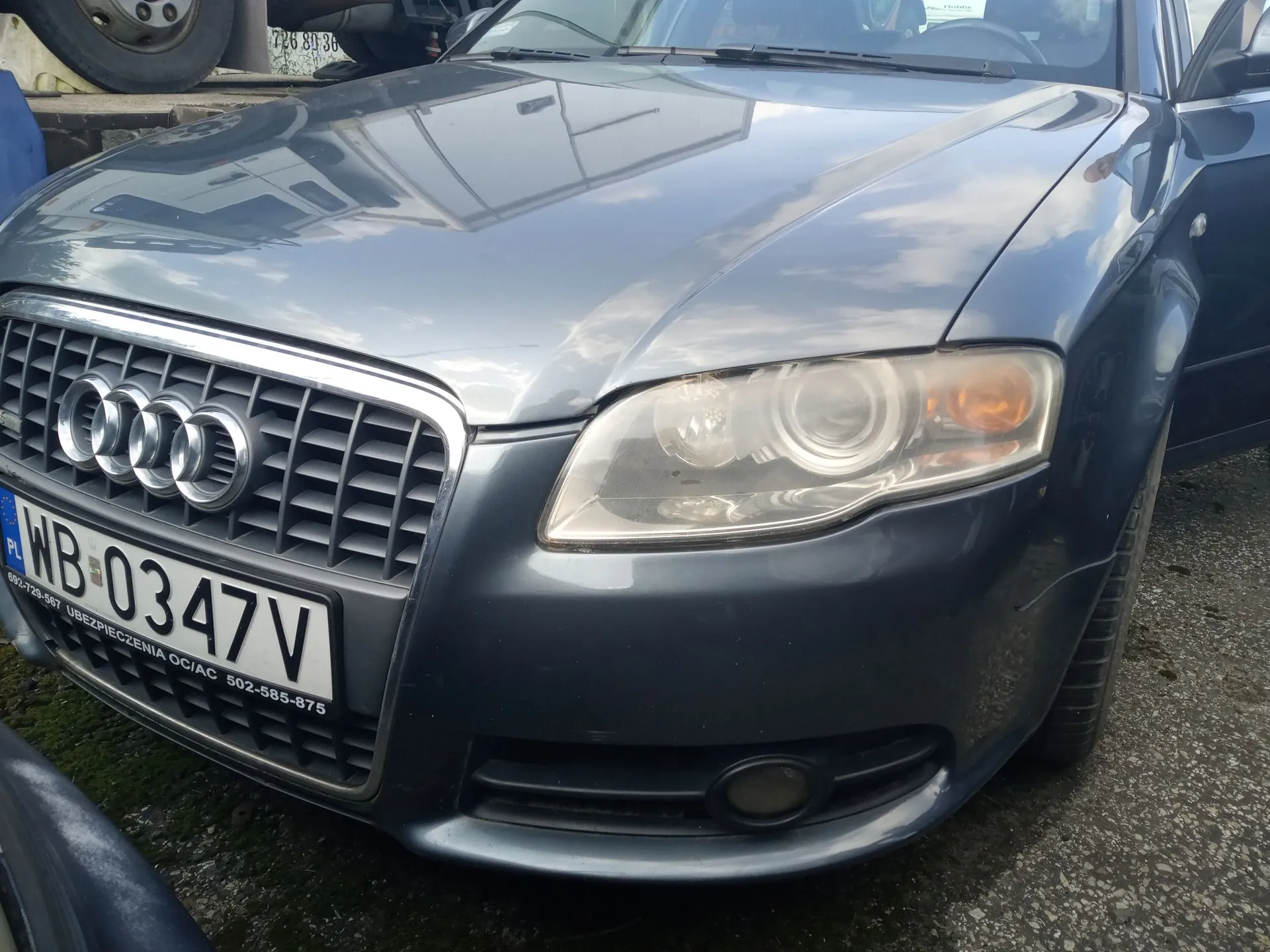 Zbliżenie na przód szarego samochodu marki Audi. Widoczny jest charakterystyczny grill z czterema pierścieniami oraz polska tablica rejestracyjna o numerze WB 0347V. Poniżej numeru rejestracyjnego znajduje się napis: '692-729-567 UBEZPIECZENIA OC/AC 502-585-875'. Widoczny jest prawy reflektor oraz fragment przedniego zderzaka z mgiełką.