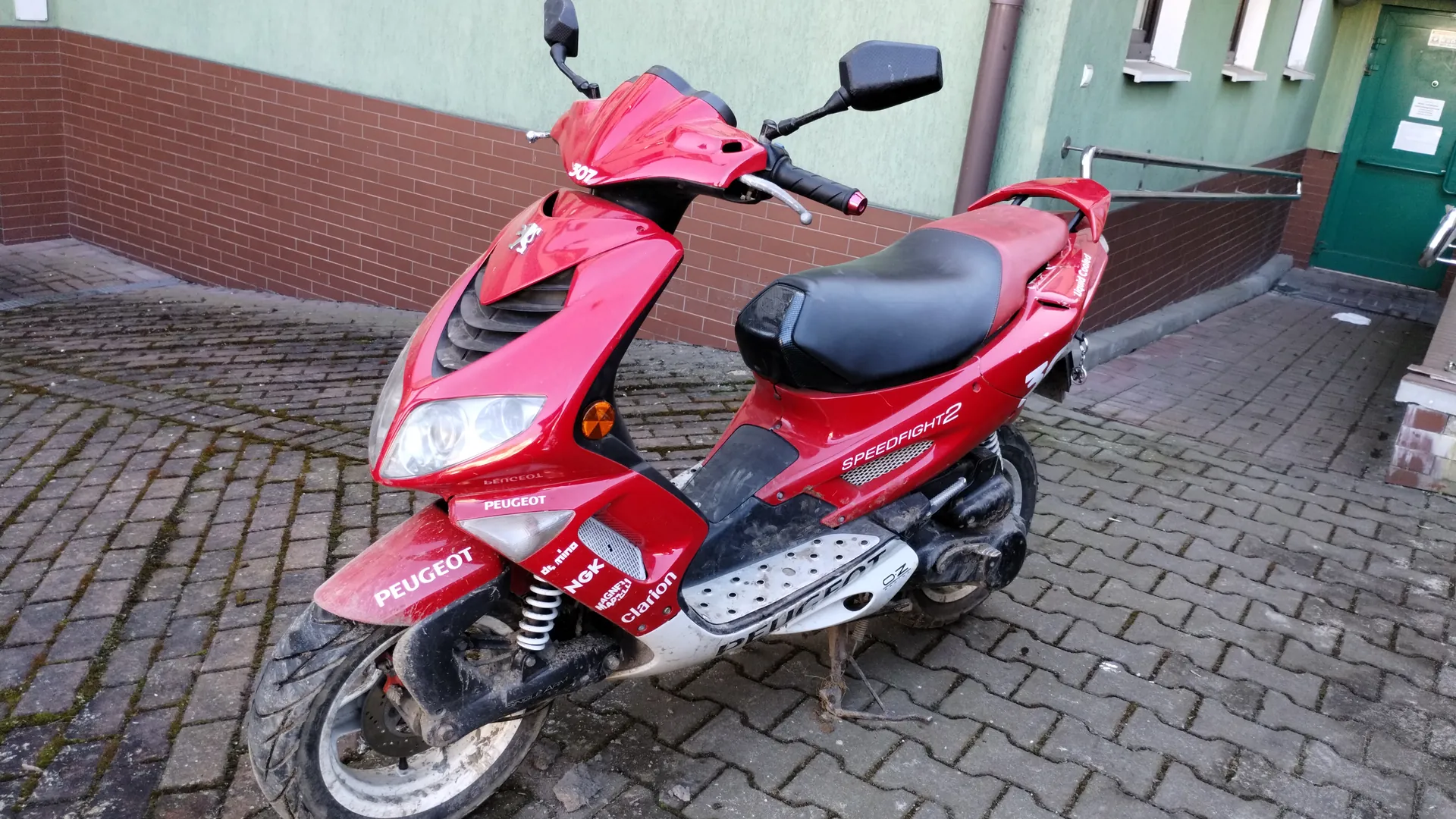 Czerwony skuter marki Peugeot model Speedfight 2 zaparkowany na brukowanej powierzchni. Skuter posiada czarne siedzenie, lusterka boczne oraz widoczne napisy: PEUGEOT na przodzie, SPEEDFIGHT 2 na boku, NGK i Clarion na czerwonej części przedniej. W tle zielono-brązowa ściana budynku z cegły, zielone drzwi i okna. Podłoga wyłożona kamiennymi płytami z widocznymi śladami mchu.