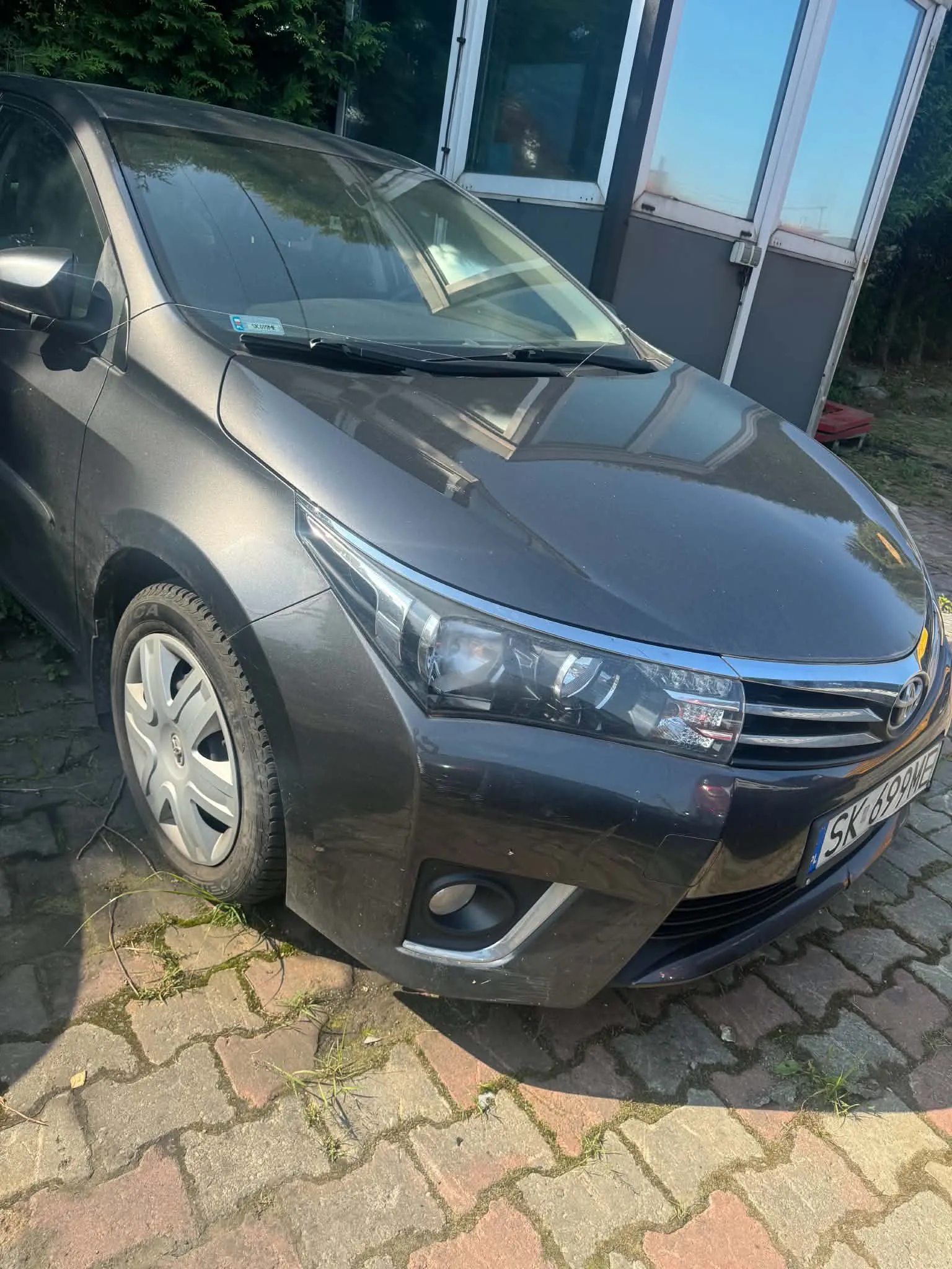 Szary samochód osobowy marki Toyota zaparkowany na kostce brukowej przed budynkiem. Widoczny jest przód pojazdu z tablicą rejestracyjną o numerze SK 699NE.