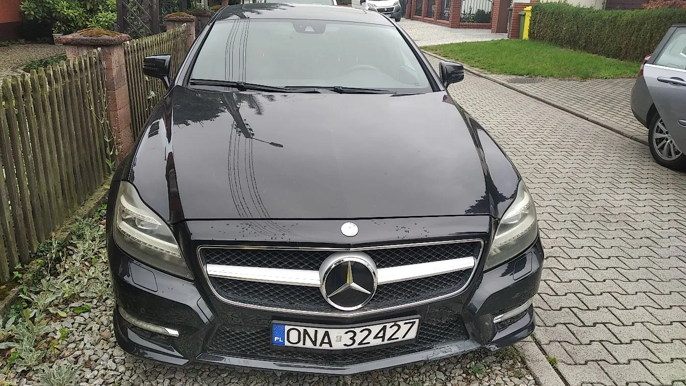 Czarny samochód marki Mercedes-Benz zaparkowany na podjeździe z kostki brukowej. Widoczny jest przód pojazdu z charakterystycznym logo gwiazdy na grillu oraz polska tablica rejestracyjna o numerze ONA 32427. W tle znajduje się drewniany płot, zieleń oraz fragment innego samochodu po prawej stronie.