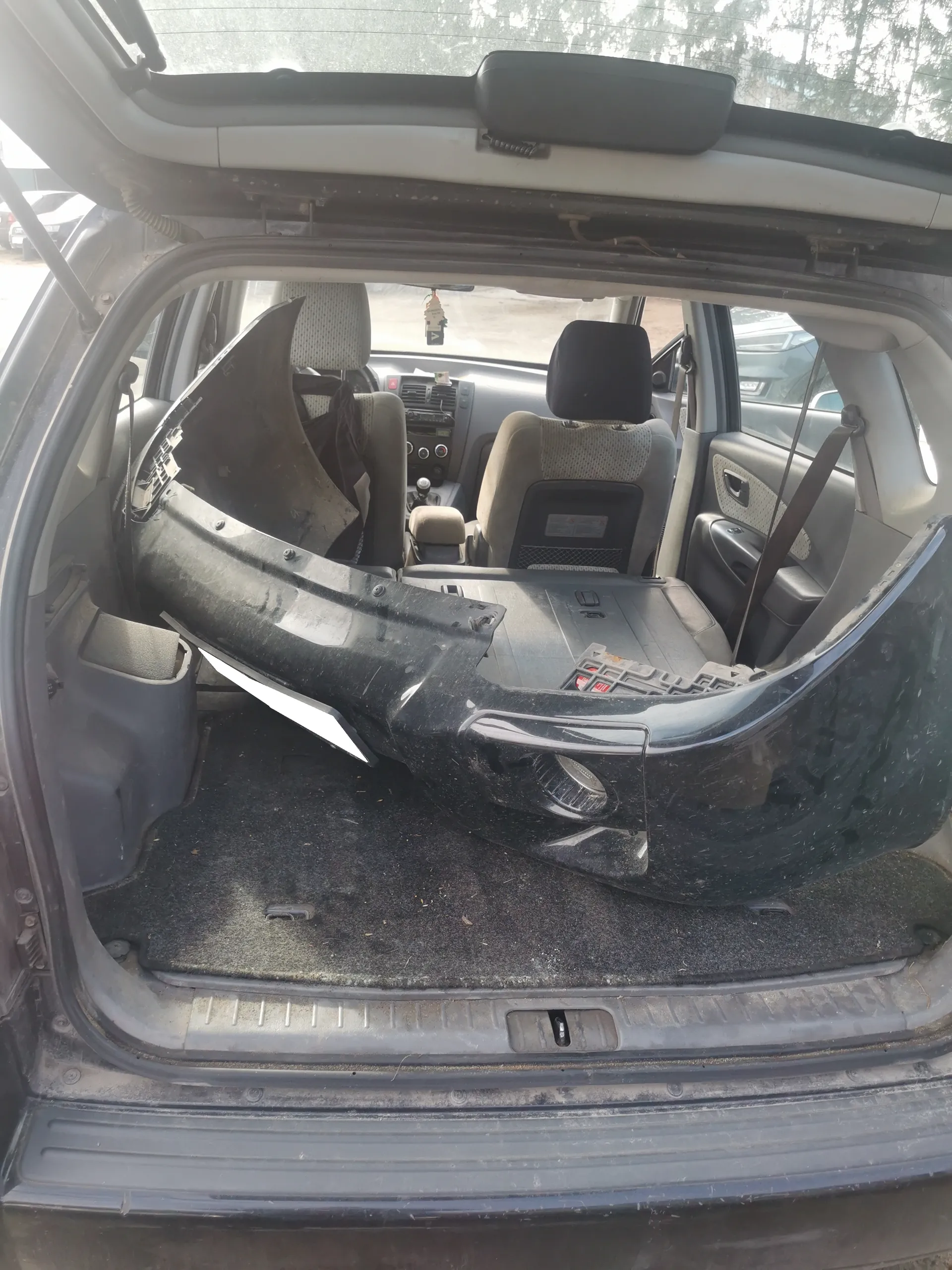 Wnętrze samochodu widziane od tyłu z otwartym bagażnikiem. W bagażniku leży uszkodzony czarny plastikowy element karoserii. Widoczne są przednie siedzenia, deska rozdzielcza z przyciskami, kierownica oraz wiszący na lusterku wstecznym przedmiot. Podłoga bagażnika jest zabrudzona. W tle przez przednią szybę widać drzewa i inne pojazdy.