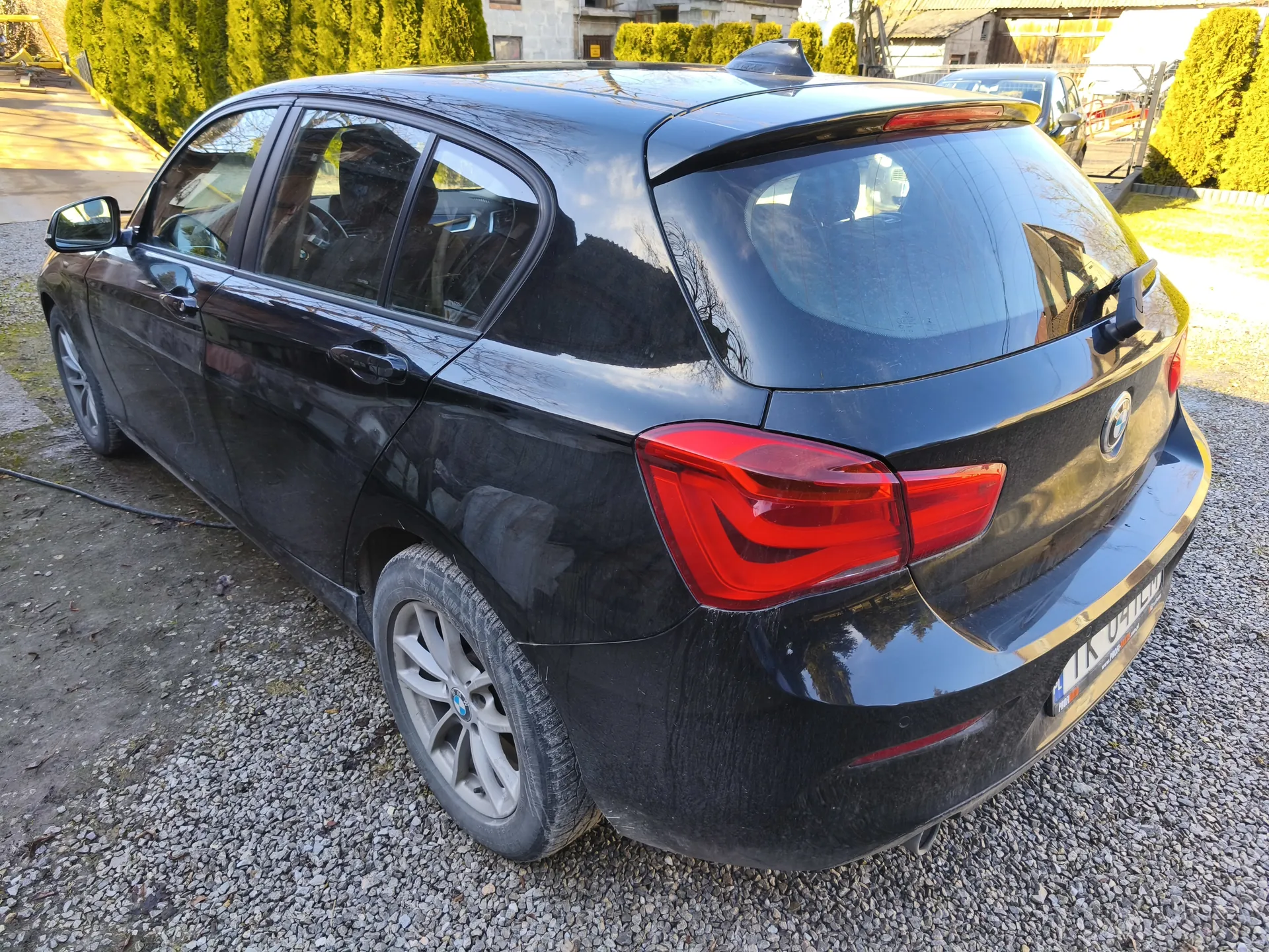 Czarny samochód osobowy marki BMW typu hatchback widziany od tyłu i boku, zaparkowany na podłożu z żwiru. Widoczne są czerwone światła tylnego, tablica rejestracyjna z napisem TK 041 EW, felgi aluminiowe oraz odbicia w szybach. W tle znajdują się inne pojazdy, zielone krzewy, budynki i fragment drogi.