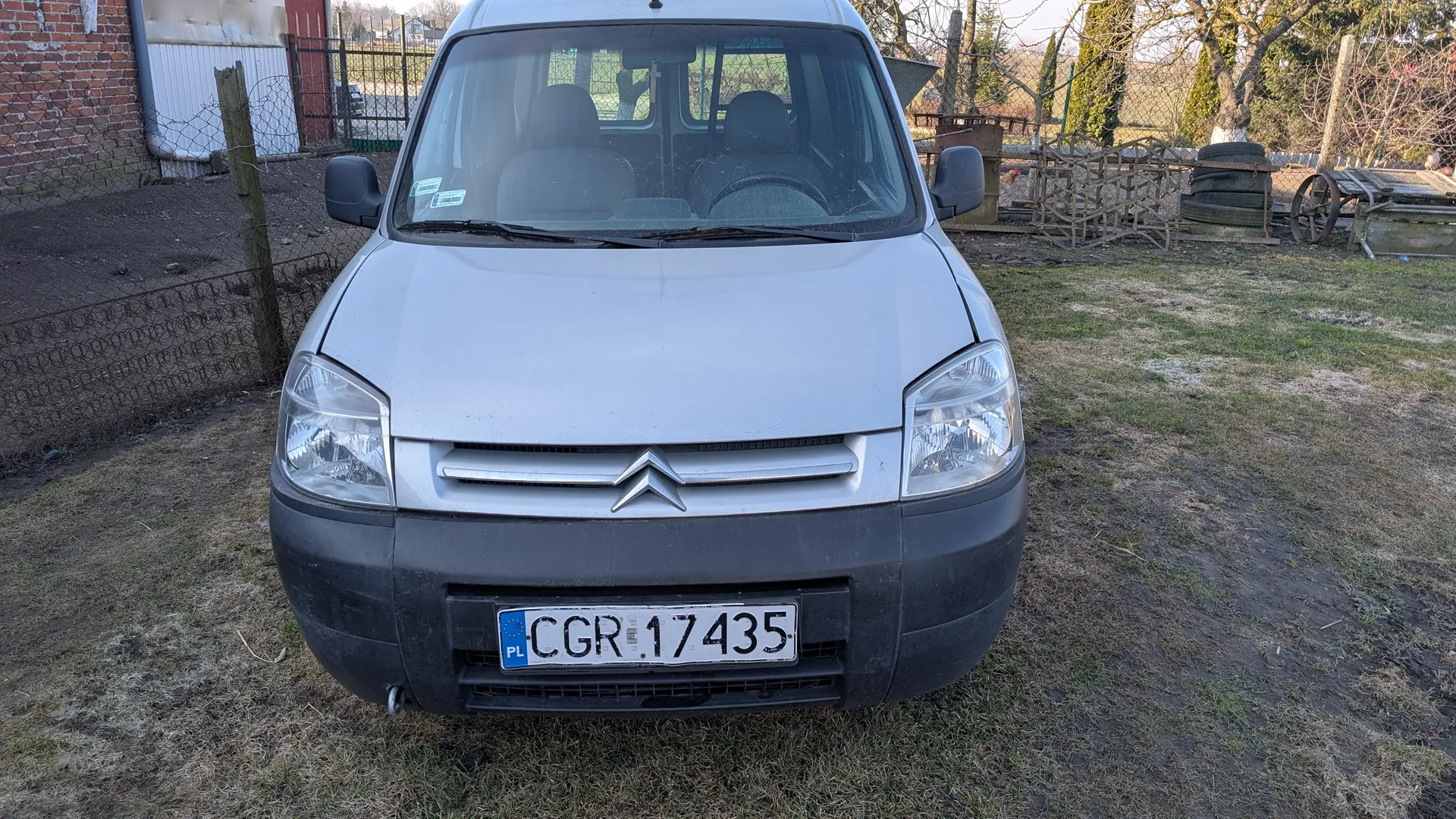 Przedni widok srebrnego samochodu dostawczego marki Citroën z tablicą rejestracyjną PL CGR 17435. Samochód stoi na trawiastym i częściowo zarośniętym błotem terenie. W tle widoczne są ceglany budynek, siatka drutu kolczastego, stos opon, drewniana konstrukcja oraz suche drzewa. Na przedniej szybie widoczne są dwa naklejki. Oświetlenie wskazuje na słoneczny dzień.
