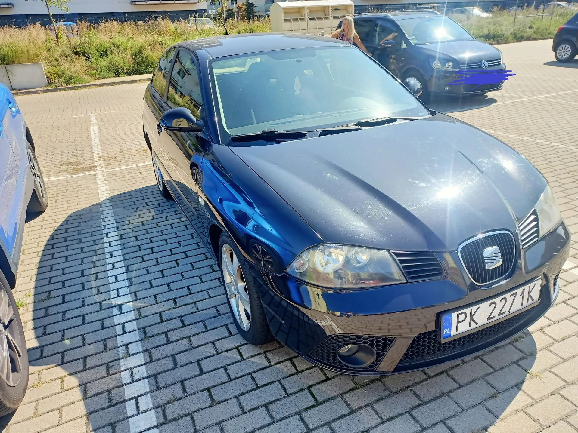 Ciemnoniebieski samochód marki Seat zaparkowany na kostce brukowej. Widoczna jest przednia część pojazdu z polską tablicą rejestracyjną o numerze PK 2271K. W tle znajduje się inny, ciemny samochód oraz fragmenty zieleni i budynków.