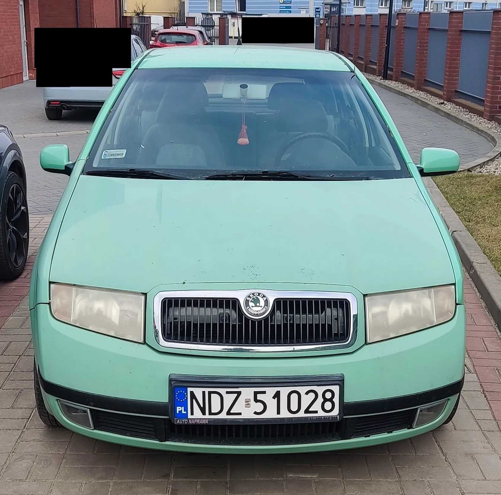 Przedni widok jasnozielonego samochodu marki Škoda zaparkowanego na brukowanej powierzchni. Na masce widoczny jest emblemat marki, a na tablicy rejestracyjnej napis "NDZ 51028" z niebieskim pasem po lewej stronie zawierającym literę "PL" oraz napisem "AUTO NAPRAWA" na dole. W tle znajdują się inne pojazdy (czerwony i srebrny samochód), ceglane budynki, ogrodzenie z cegieł i niebieskich paneli oraz trawnik. Na przedniej szybie widoczny jest naklejony znaczek, a wewnątrz samochodu wisząca ozdoba.