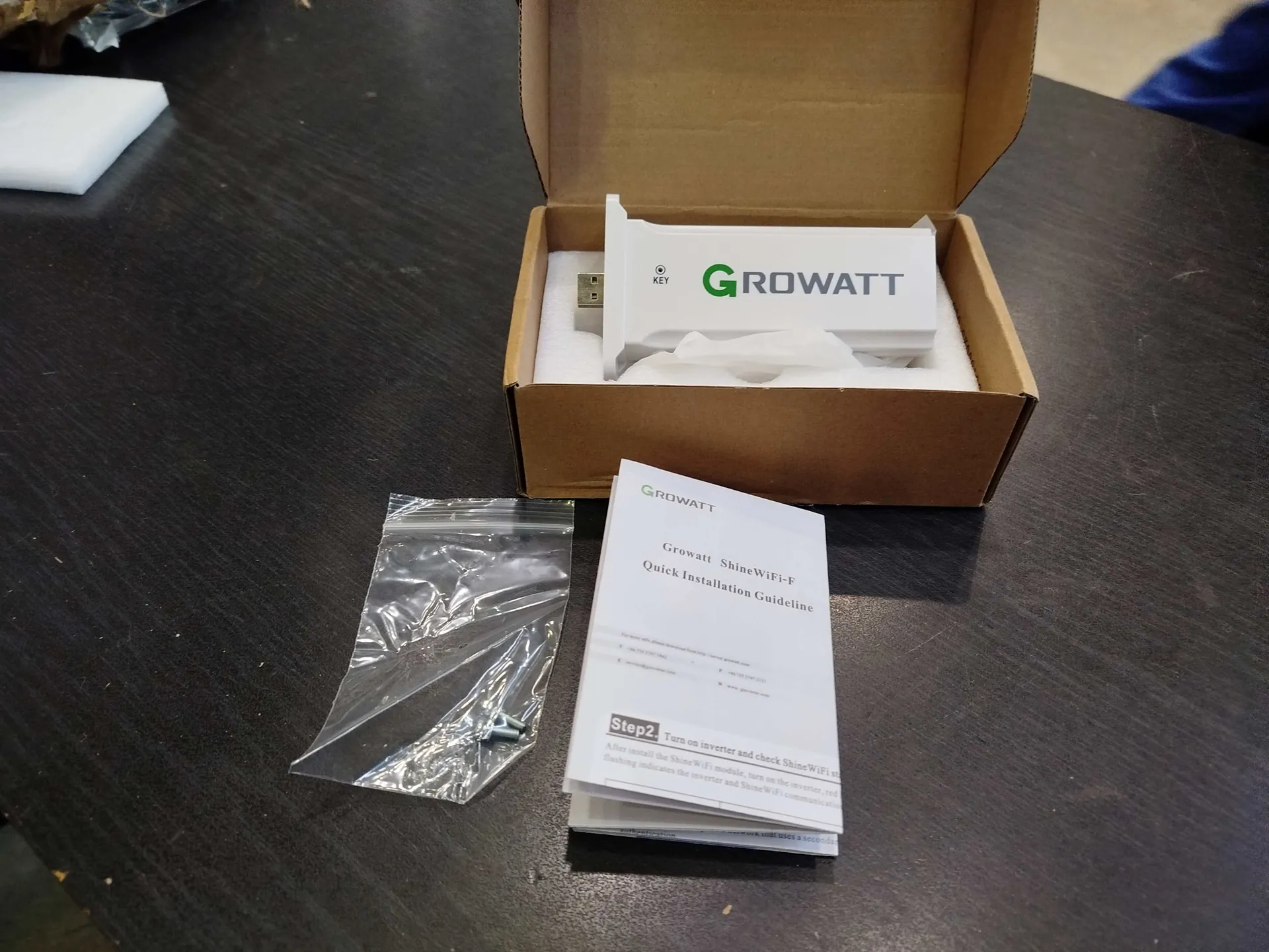 Otwarte brązowe pudełko kartonowe na ciemnym stole. Wewnątrz pudełka białe urządzenie z logo "GROWATT" (zielona litera "G" i czarne litery "ROWATT"), widoczne złącze USB. Przy pudełku leży przezroczysta torebka z małymi częściami metalowymi oraz dokument zatytułowany "Growatt ShineWiFi-F Quick Installation Guideline", na którym widoczny jest krok "Step2. Turn on inverter and check Shine WiFi...". W tle fragment białej pianki i niebieskiego przedmiotu.