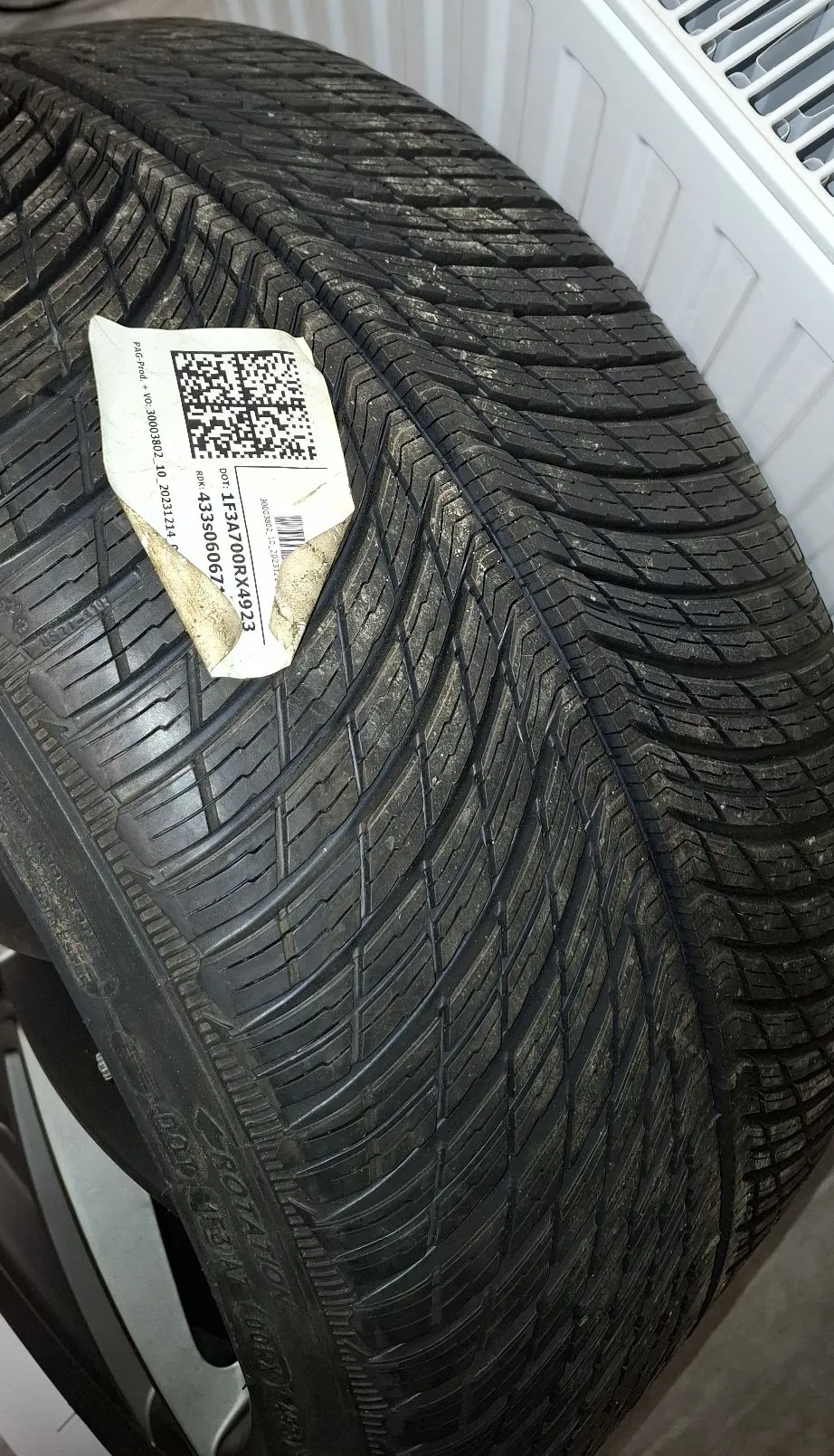 Zbliżenie na bieżnik czarnej opony samochodowej. Na boku opony przyklejona jest biała, lekko pognieciona etykieta z kodem QR oraz tekstem: 'P&R pod. + VO: 3003802 10. 20231214 0', 'SN: 4335060067*' oraz 'DOT: 1F34700RX4923'.
