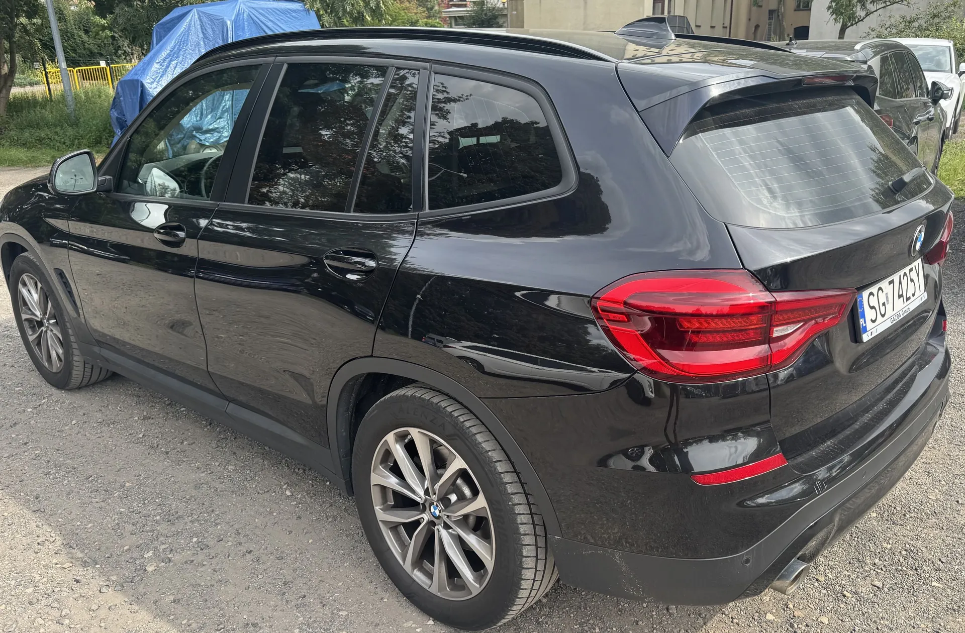 Czarny samochód marki BMW typu SUV zaparkowany na żwirowym podłożu, widoczny z tyłu i z boku. Pojazd ma przyciemniane szyby, srebrne felgi aluminiowe oraz tablicę rejestracyjną o numerze SG 7425Y. W tle widoczna jest niebieska plandeka oraz fragmenty zieleni i innego zaparkowanego samochodu.