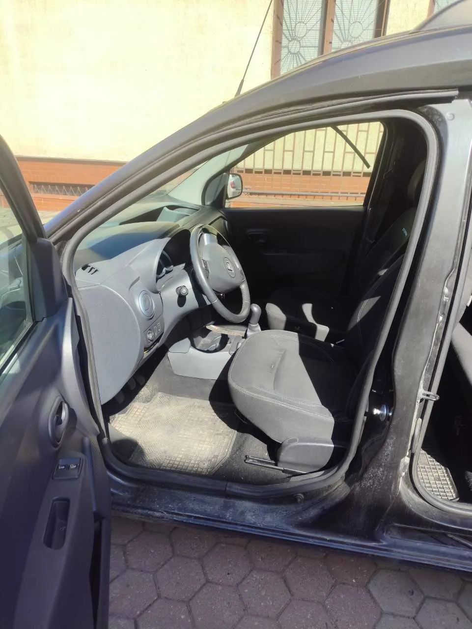 Wnętrze samochodu z otwartą lewą przodem drzwiami. Widoczne są kierownica, deska rozdzielcza w kolorze szarym, czarne i szare siedzenia oraz podłoga z matą. Poza samochodem widać brukowaną powierzchnię z sześciokątnymi płytami oraz fragment budynku z ceglaną ścianą i oknem z ozdobną kratą. Drzwi pasażerskie są zamknięte, a zewnętrzne lusterko boczne widoczne po lewej stronie.
