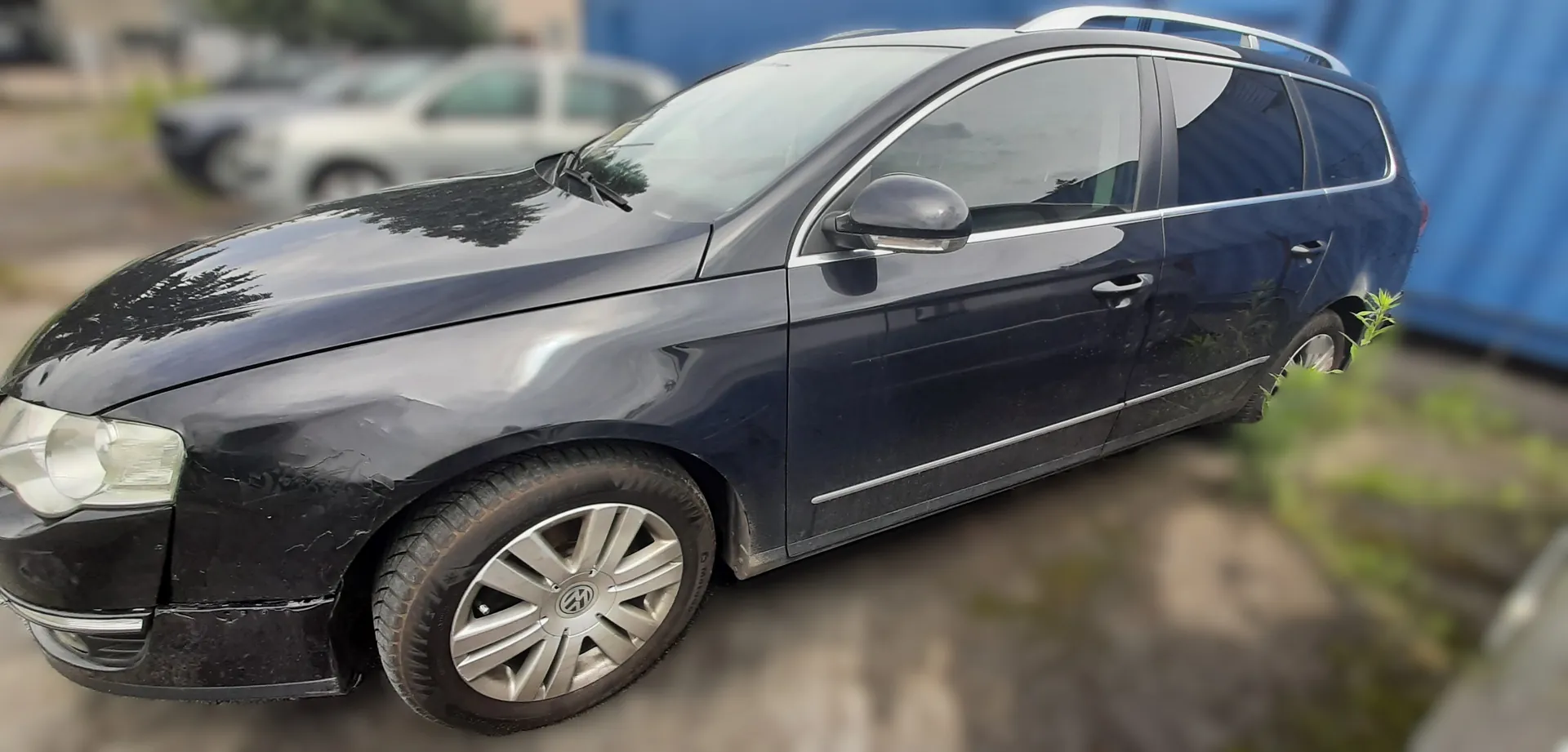 Ciemnoszary samochód osobowy typu kombi marki Volkswagen z widocznymi uszkodzeniami na przedniej lewej stronie, w tym zadrapaniami i wgnieceniami. Samochód posiada srebrne felgi z logo VW. W tle znajdują się inne pojazdy, zieleń oraz niebieska konstrukcja. Zdjęcie wykonane na zewnątrz, prawdopodobnie na parkingu.