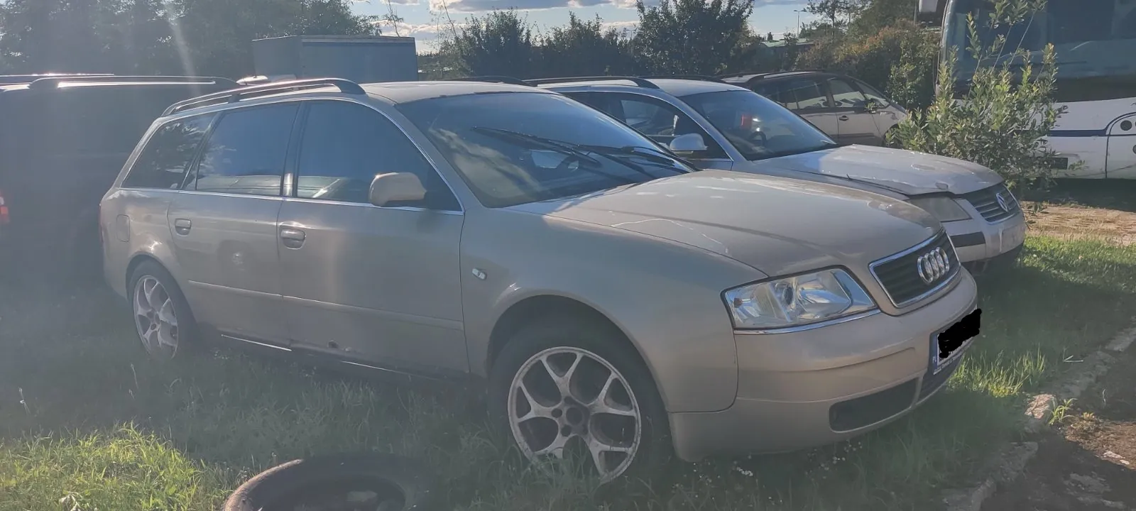 Jasnoszary samochód marki Audi w nadwoziu kombi, zaparkowany na trawie obok innych pojazdów. Auto jest widoczne z przodu i z boku, ma srebrne felgi aluminiowe. W tle widać biały samochód osobowy oraz fragment białego autobusu.