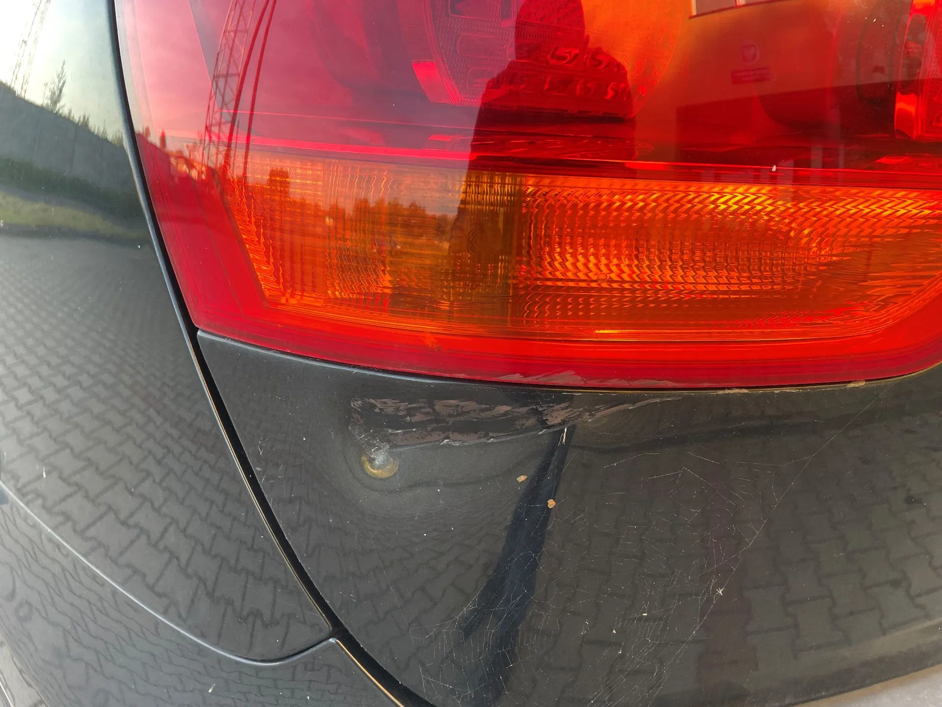 Zbliżenie na tylną lampę samochodu w kolorze czerwonym i pomarańczowym oraz przylegający do niej czarny element nadwozia, który posiada widoczne zarysowania i wgniecenie.