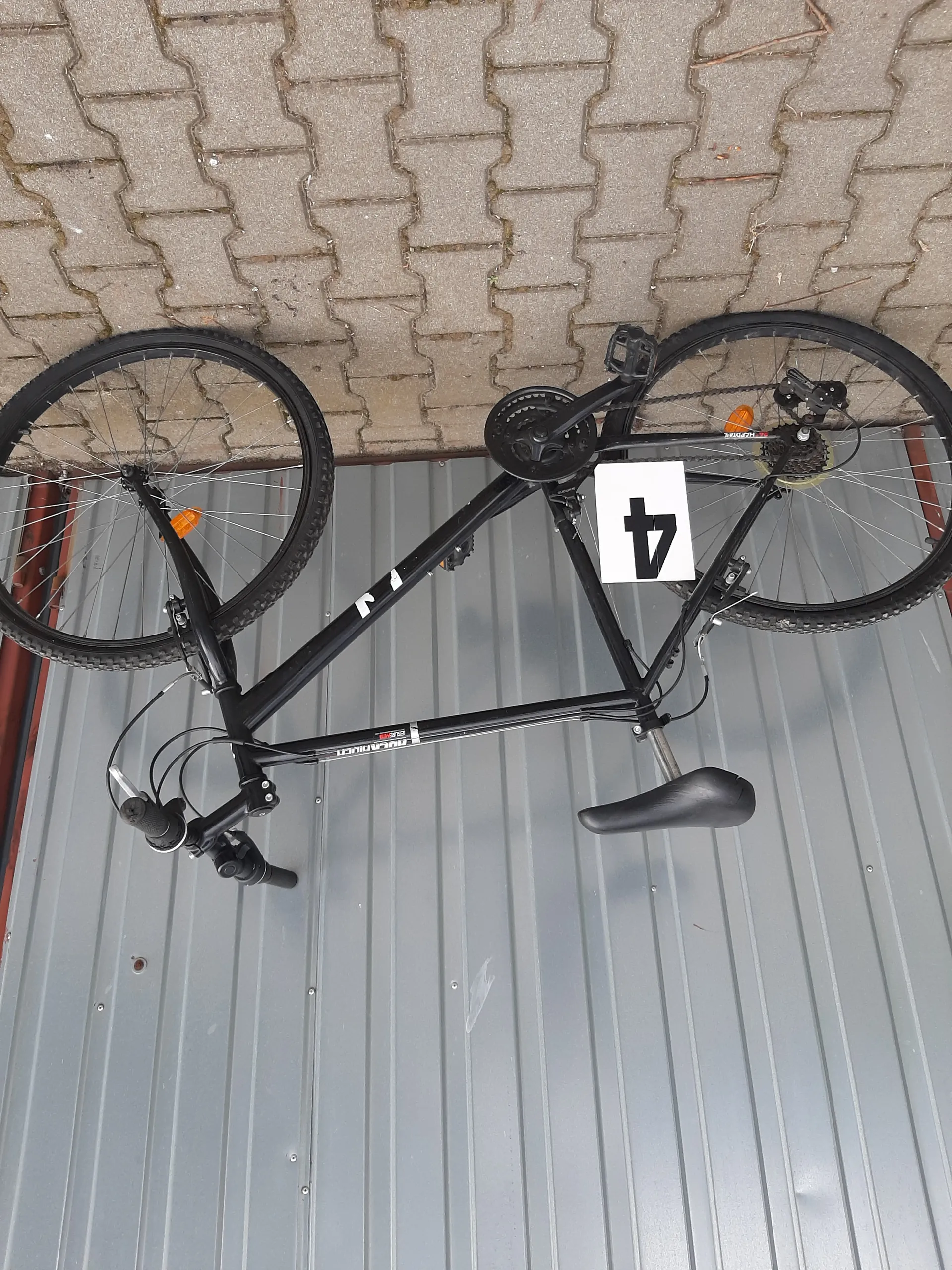 Czarny rower oparty o szarą metalową ścianę z poziomymi listwami i brukowaną powierzchnię. Na ramie roweru umieszczony biały znak z czarną cyfrą 4. Rower posiada czarne koła, czarne siedzisko oraz kierownicę z czarnymi uchwytami. W tle metalowa ściana i bruk w kształcie ząbków.