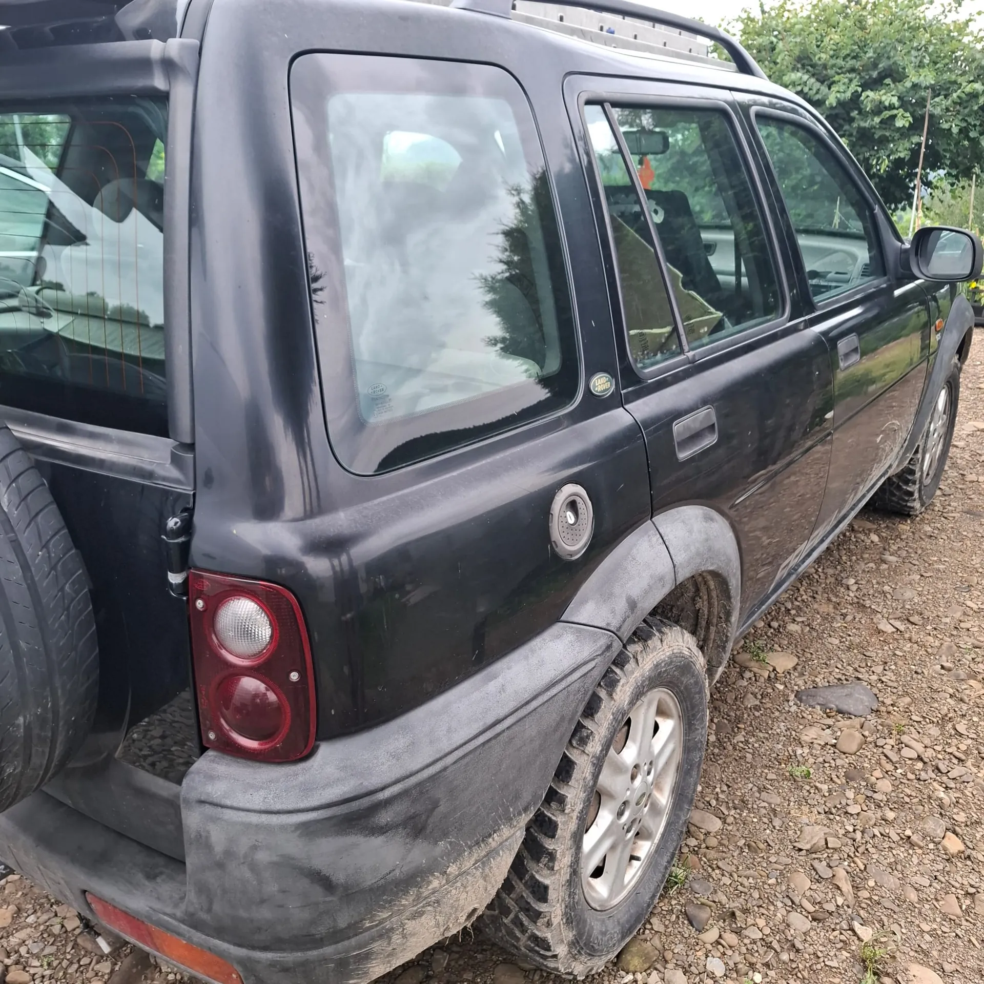 Zbliżenie na tylną i boczną część czarnego samochodu terenowego typu SUV. Pojazd jest zakurzony i stoi na kamienistym podłożu. Widoczna jest tylna szyba, boczne okna, koło zapasowe zamontowane na klapie oraz tylna lampa z czerwonymi kloszami.