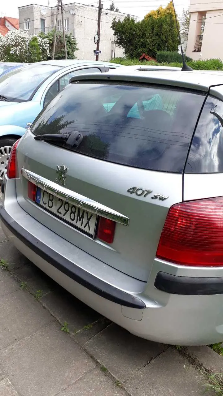 Tył srebrnego samochodu Peugeot 407 SW z tablicą rejestracyjną CB 298MX z kodem kraju PL. Samochód zaparkowany na chodniku z widocznymi szczelinami i trawą. W tle inne pojazdy, budynki mieszkalne, drzewa, krzewy oraz słup telekomunikacyjny. Niebo częściowo zachmurzone.